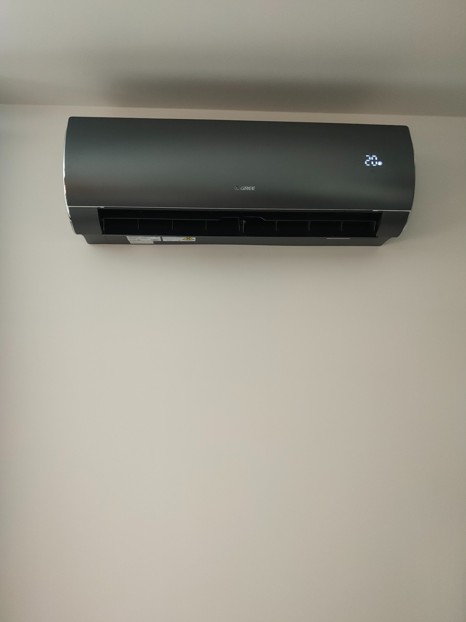 Czarny klimatyzator marki Gree zamontowany na jasnej ścianie, wyświetlający temperaturę 20 stopni Celsjusza.