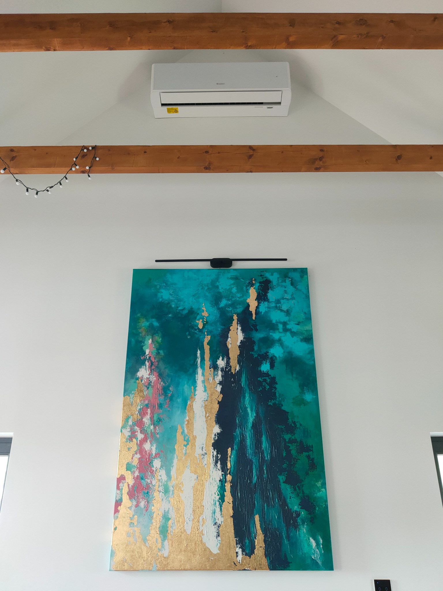 Biała klimatyzacja zamontowana pod drewnianą belką stropową w pomieszczeniu z abstrakcyjnym obrazem w odcieniach turkusu, złota i różu.