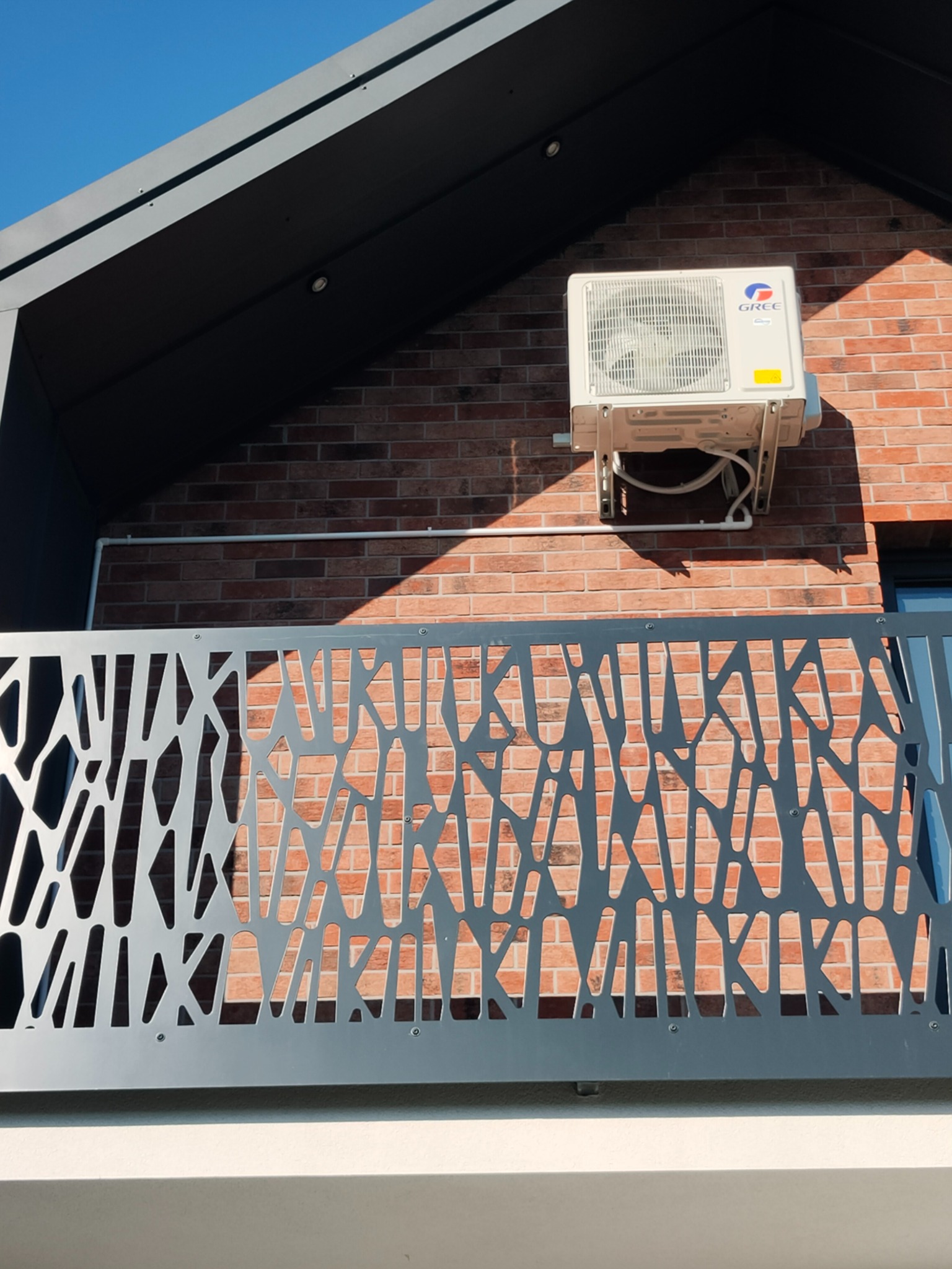 Zewnętrzna jednostka klimatyzacji marki Gree zamontowana na ceglanej ścianie nowoczesnego budynku z ozdobną, metalową balustradą balkonu.