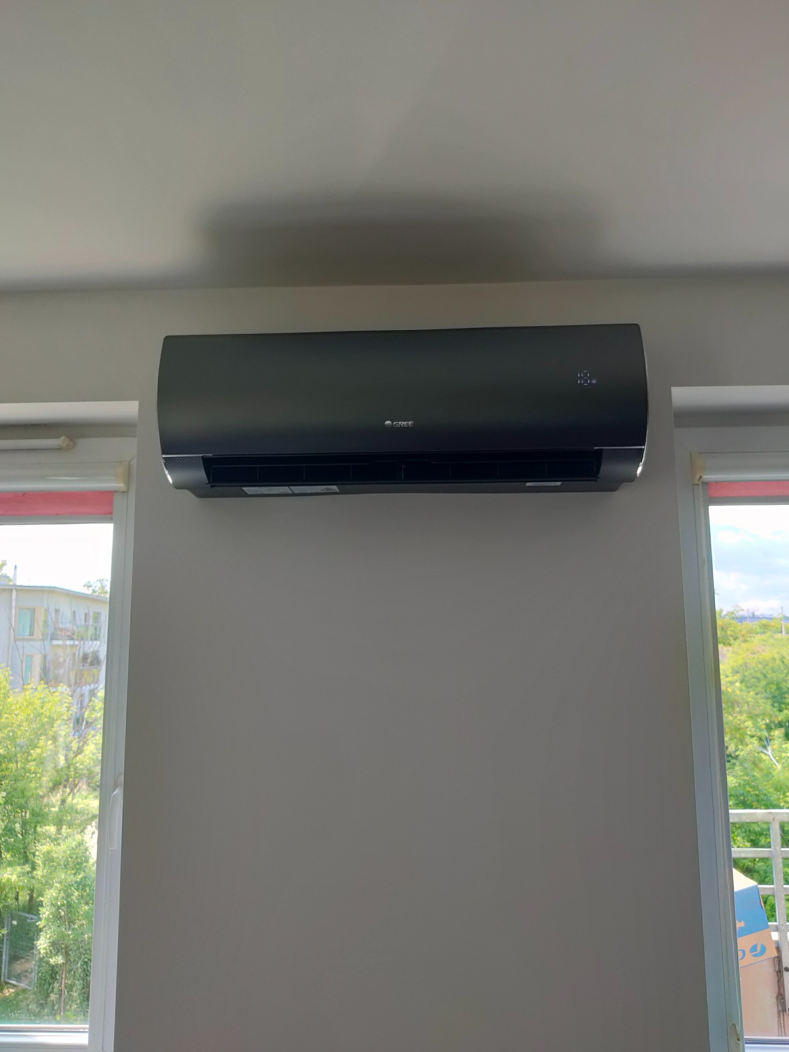 Czarny klimatyzator marki Gree zamontowany na ścianie w pomieszczeniu, widok z okna na zieleń.