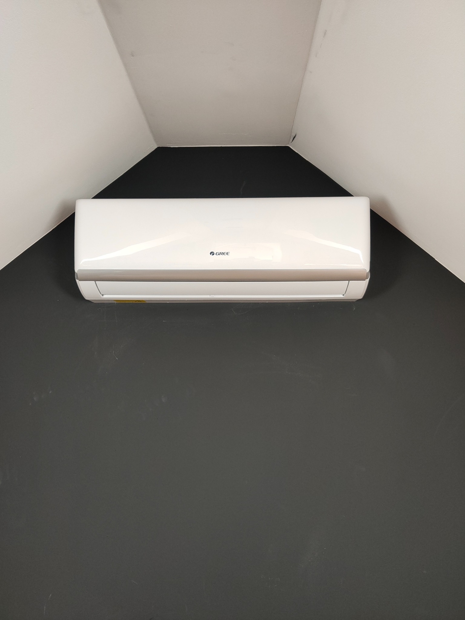 Biała jednostka klimatyzacyjna marki Gree zamontowana na ciemnoszarej ścianie pod skośnym sufitem.