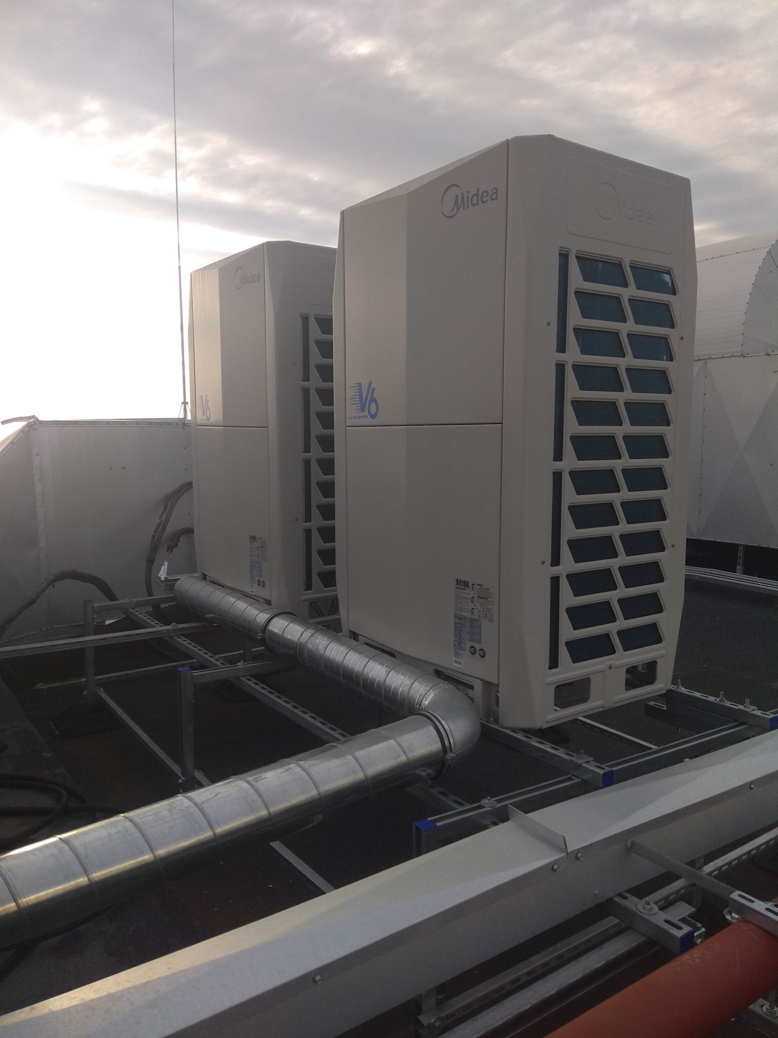 Dwa moduły zewnętrzne klimatyzacji Midea V6 na dachu budynku, połączone metalowymi rurami wentylacyjnymi, widoczne elementy konstrukcji wsporczej i fragment dachu. W tle pochmurne niebo.
