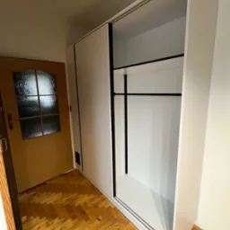 Biała szafa wnękowa z przesuwnymi drzwiami i czarnym drążkiem na ubrania, widoczna na tle drewnianej podłogi i drzwi w Gdyni.