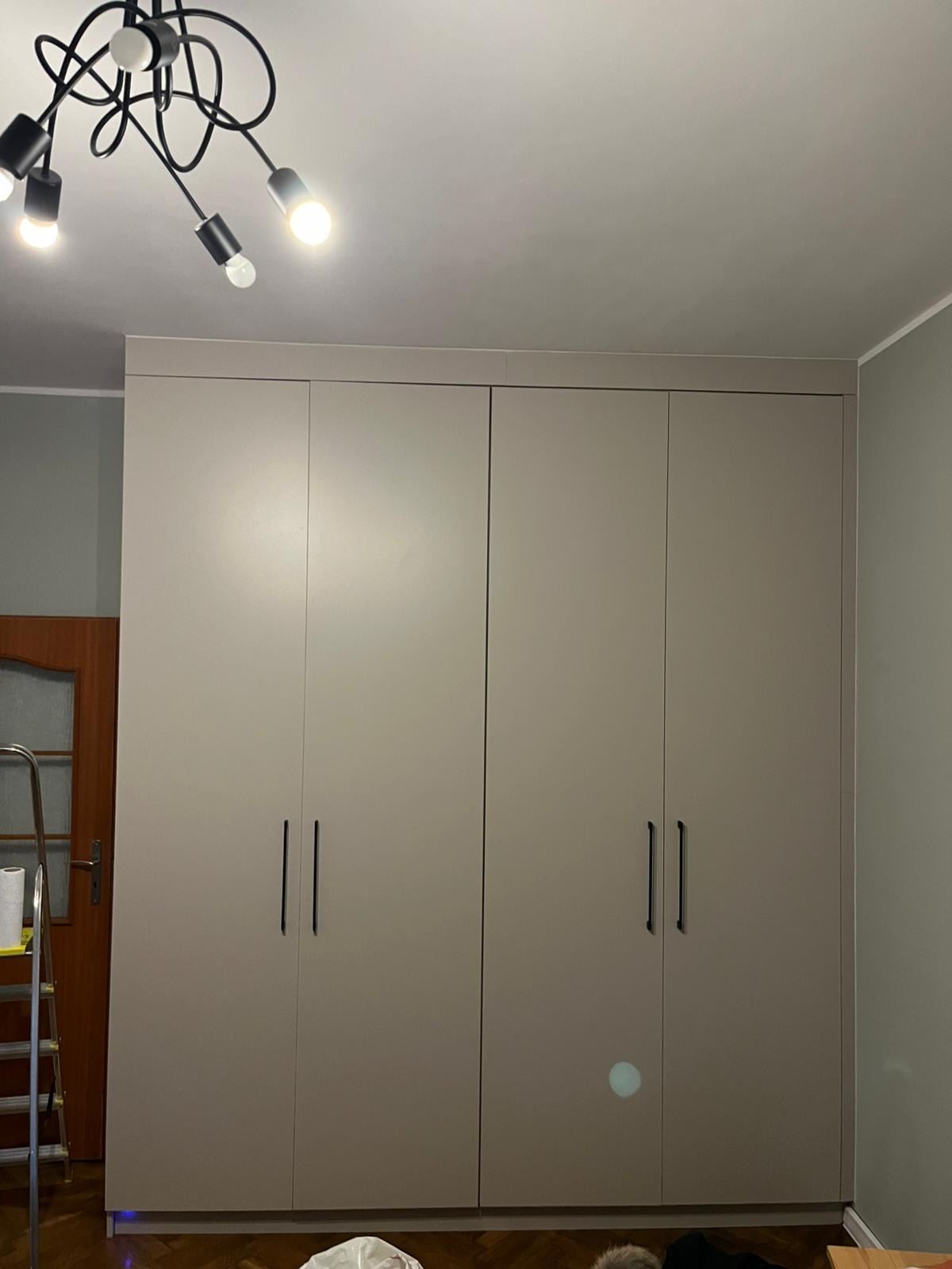 Czterodrzwiowa, beżowa szafa z minimalistycznymi, pionowymi uchwytami, zamontowana pod sufit. Widoczna drabina malarska po lewej stronie sugeruje montaż lub prace wykończeniowe.