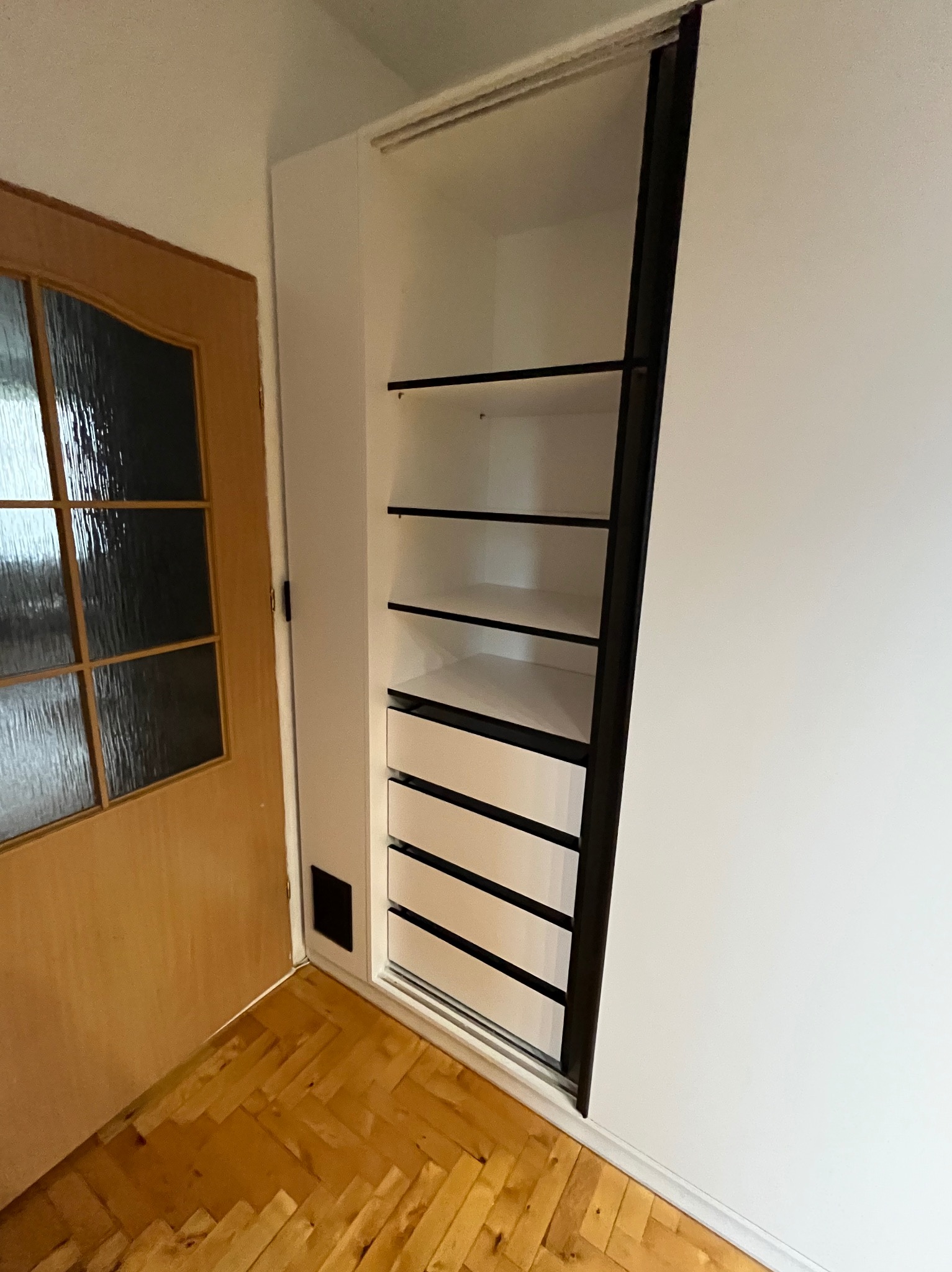 Biała szafa wnękowa z czarnymi akcentami: półki i szuflady. Widoczny fragment drewnianej podłogi parkietowej i drzwi z szybami w tle.