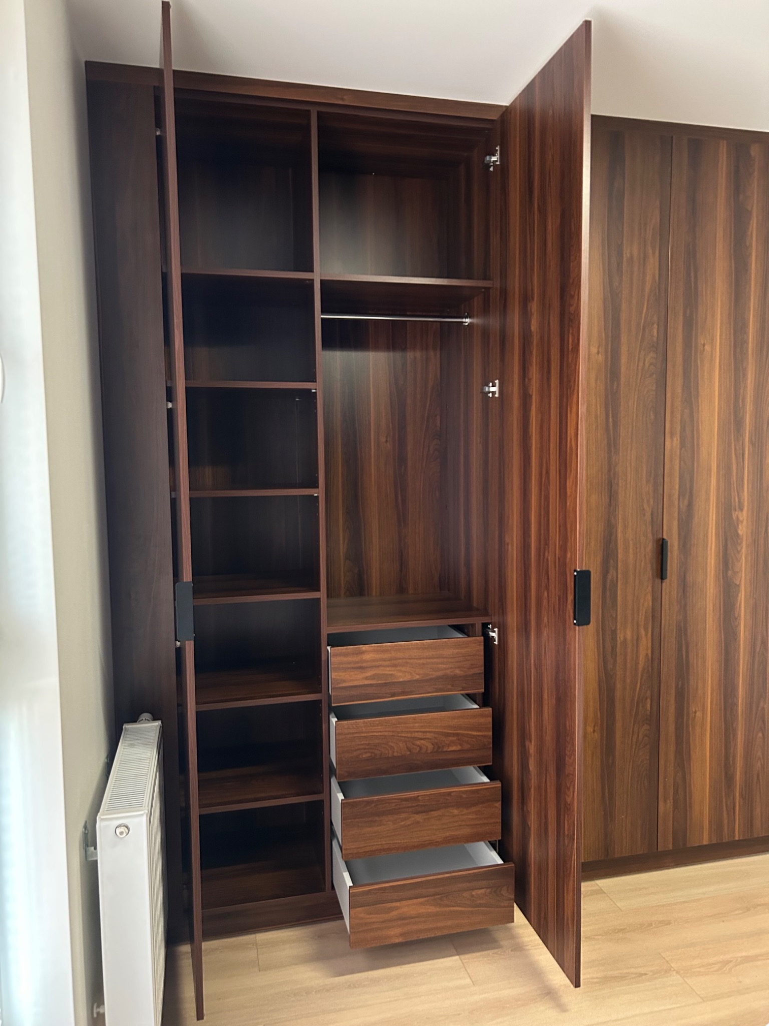Szafa wnękowa z forniru w odcieniach brązu, z otwartymi drzwiami ukazującymi półki, szuflady i drążek na ubrania. Obok widoczna kolejna szafa z zamkniętymi drzwiami.
