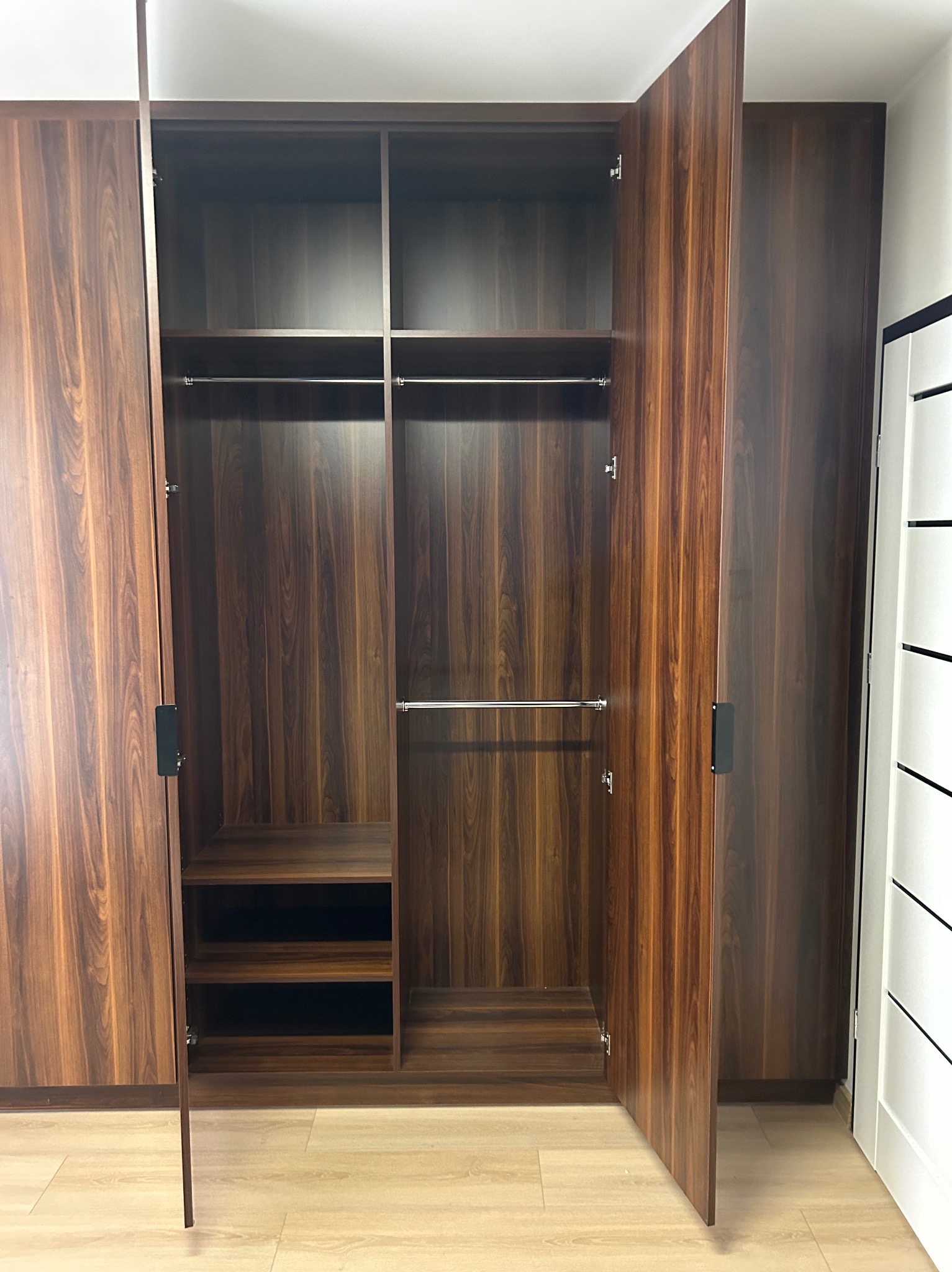 Wnętrze szafy wnękowej z otwartymi drzwiami, ukazujące półki i drążki na ubrania, wykonanej z drewna w odcieniach brązu. Widoczna podłoga i fragment białych drzwi.