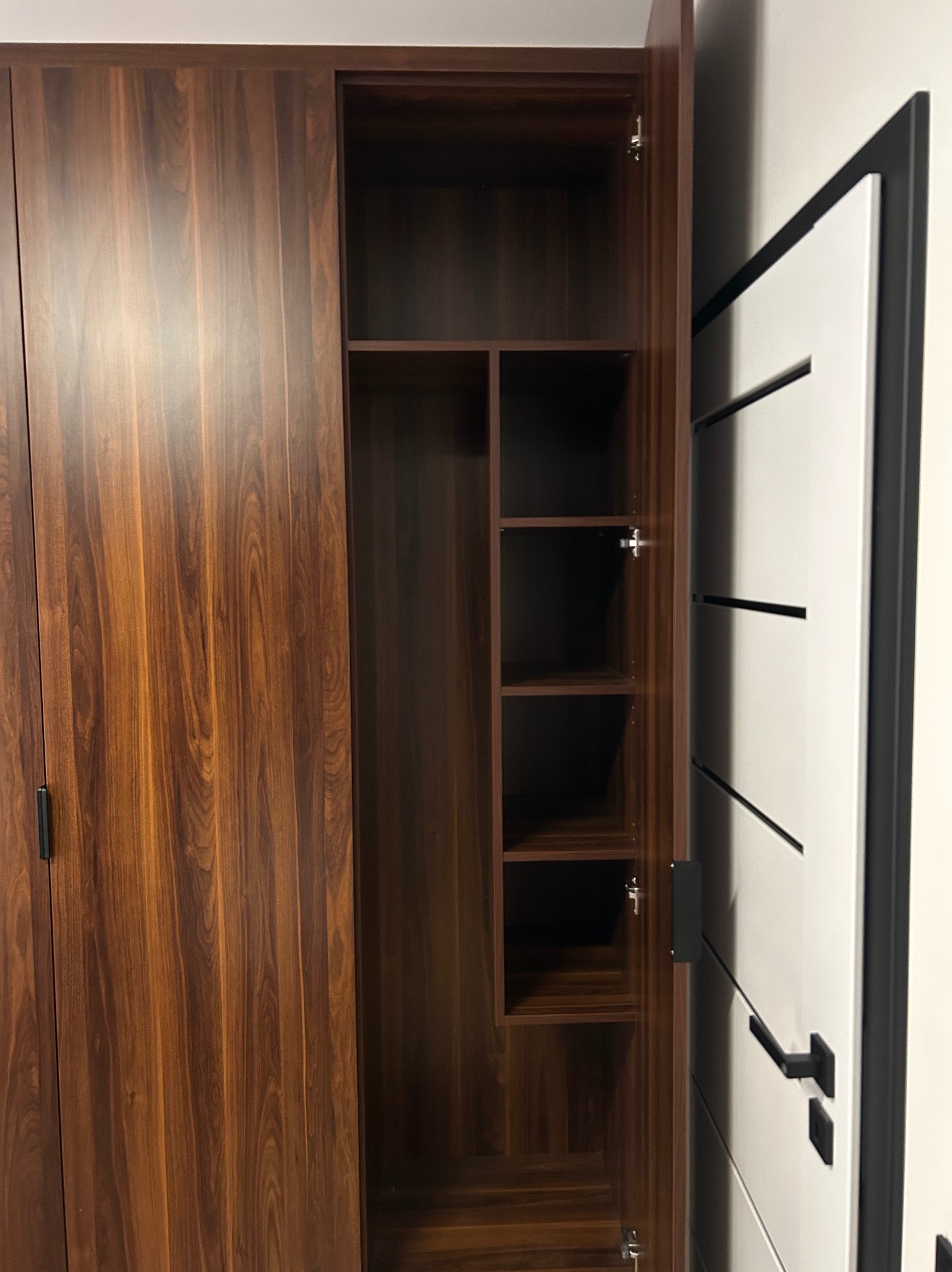 Szafa z forniru w odcieniach brązu, z otwartymi drzwiami ukazującymi półki. Obok widoczna biała, nowoczesna ściana z drzwiami z czarnymi akcentami i klamką.