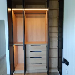 Otwarta, trzyskrzydłowa szafa z jasnego drewna z czarnymi obramowaniami i uchwytami, podświetlana od wewnątrz listwami LED, z widocznymi półkami, drążkami i szufladami.