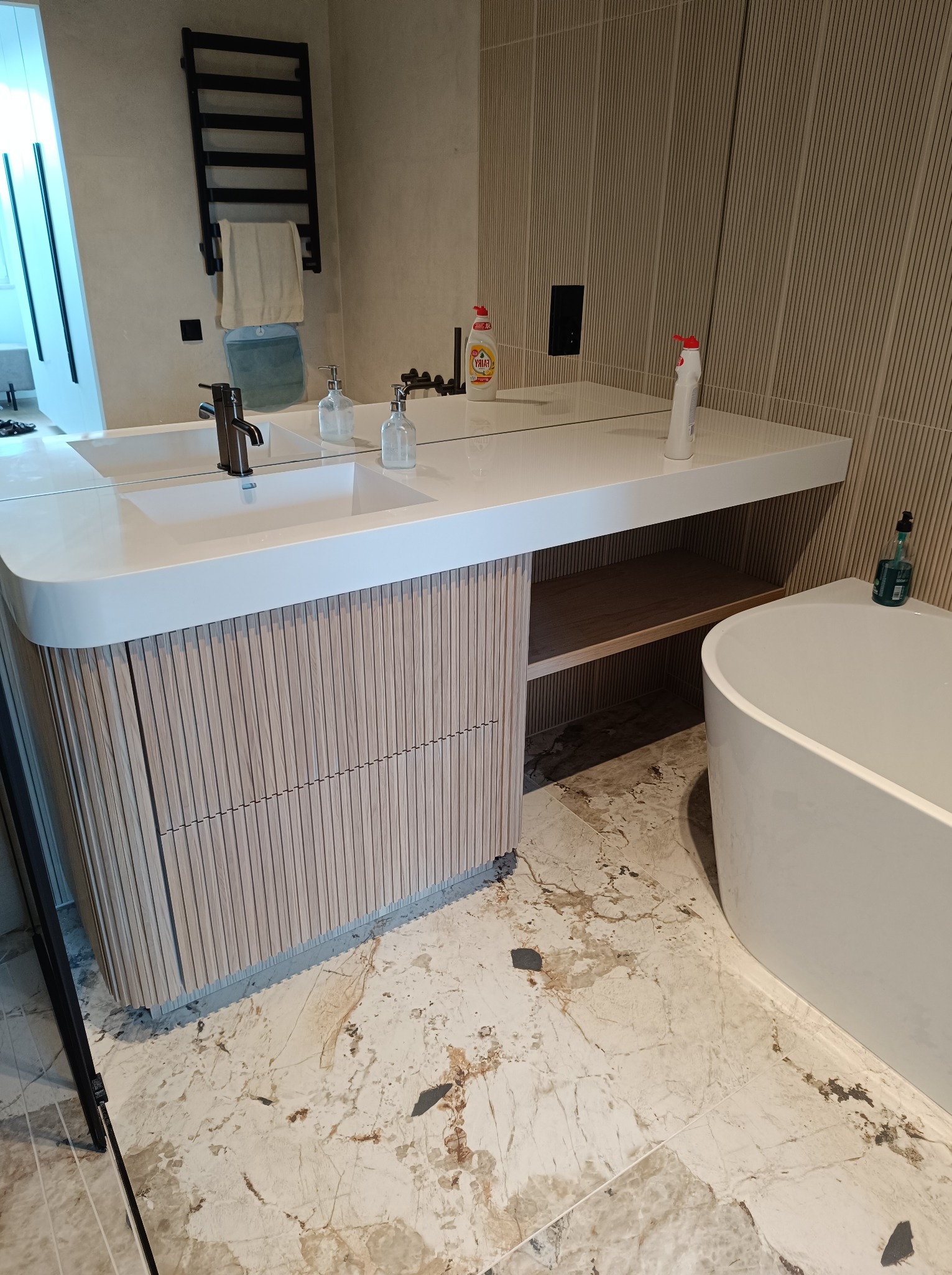 Nowoczesna łazienka z umywalką nablatową na szafce z drewnianymi lamelami, biały blat, lustro na całej długości, obok wanna wolnostojąca i grzejnik drabinkowy.