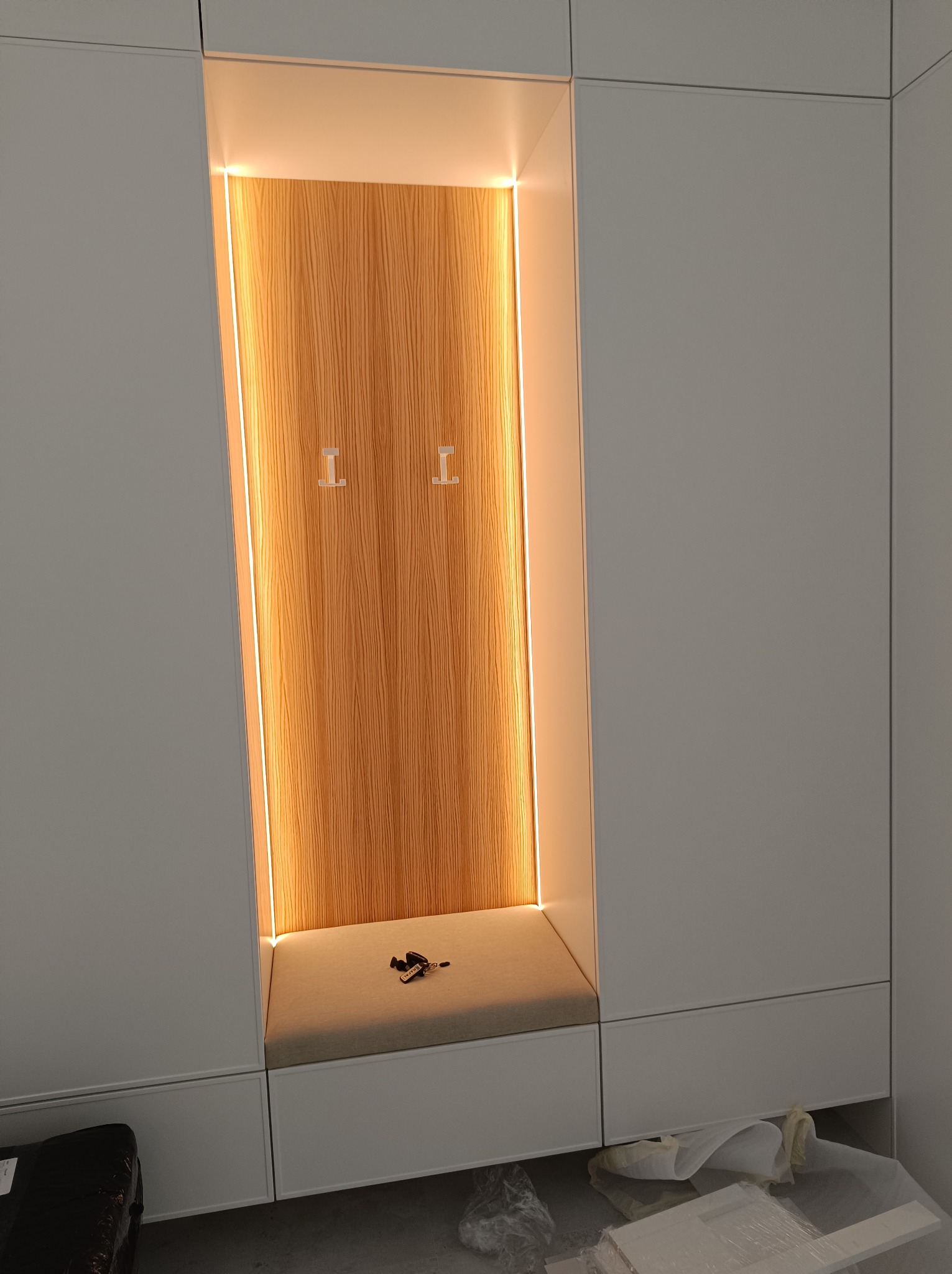 Wnęka w zabudowie meblowej z drewnianym panelem oświetlonym listwami LED, z siedziskiem i haczykami na ubrania.