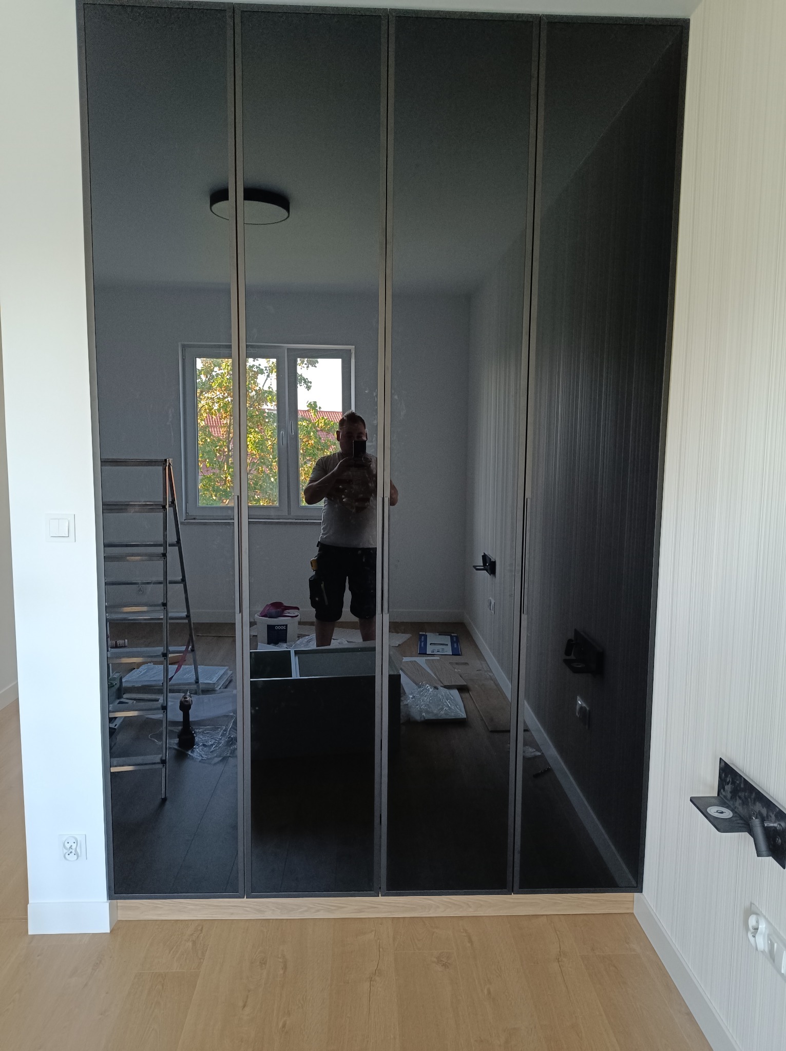 Czterodrzwiowa szafa z lustrzanymi frontami w szarej ramie, widoczne elementy montażowe i drabina w odbiciu, jasne panele na podłodze.