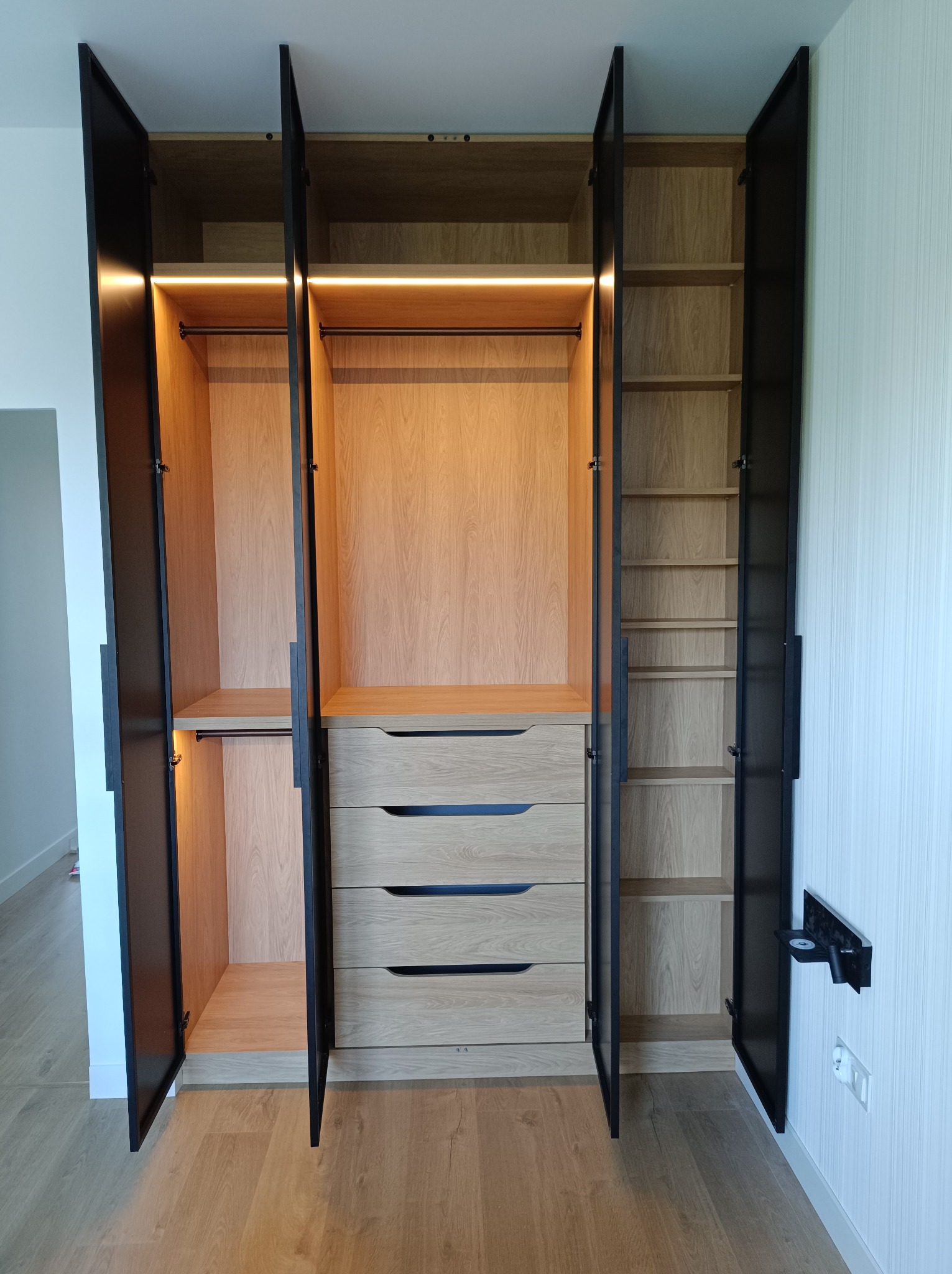 Otwarta, trzyskrzydłowa szafa z jasnego drewna z czarnymi obramowaniami i uchwytami, podświetlana od wewnątrz listwami LED, z widocznymi półkami, drążkami i szufladami.