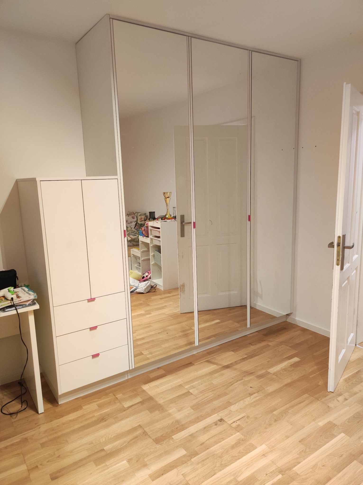 Biała szafa przesuwna z lustrzanymi drzwiami, obok mniejsza komoda z szufladami, widoczne odbicie pokoju z dziecięcymi zabawkami i otwarte drzwi.