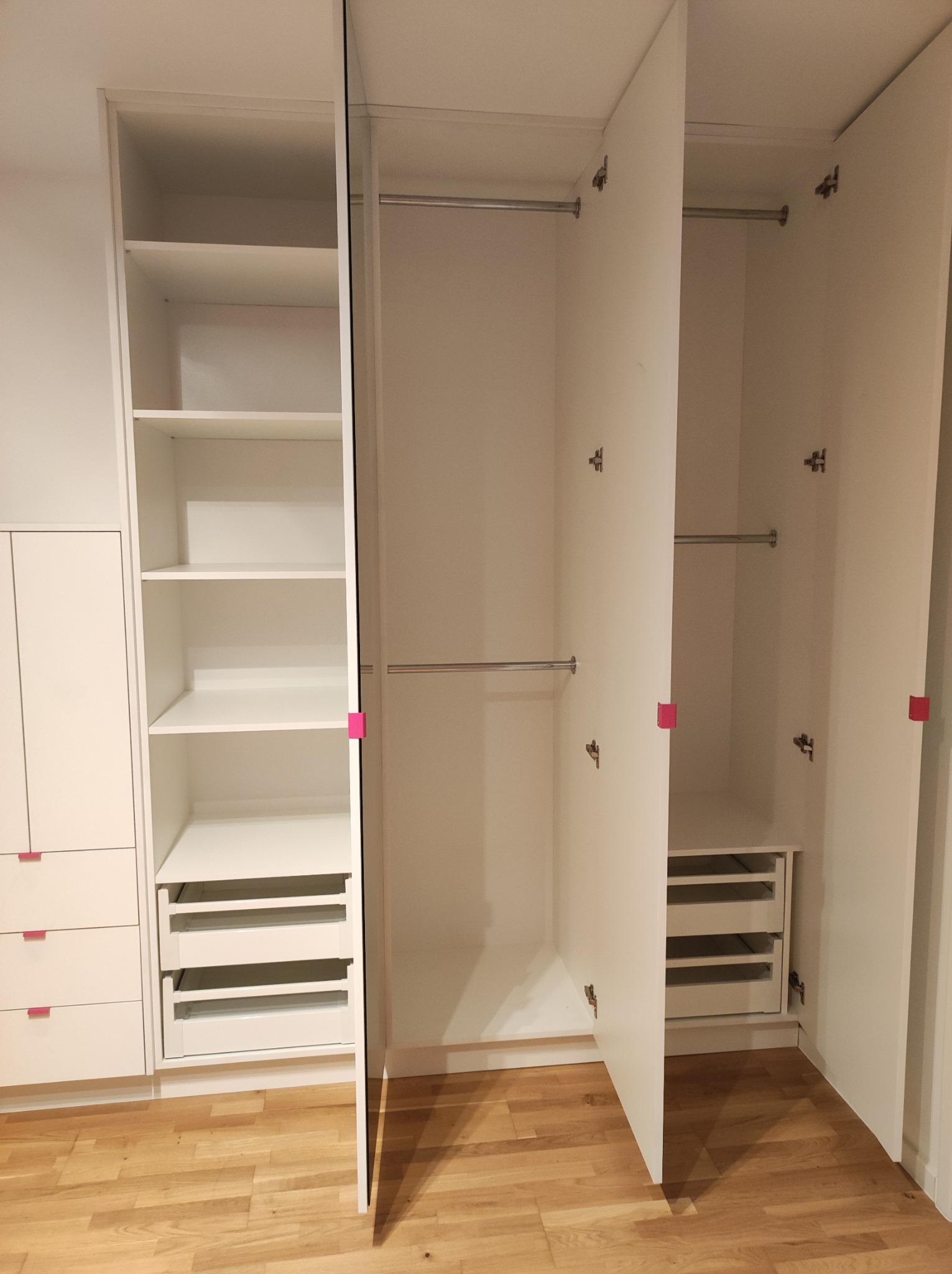 Wnętrze białej, otwartej szafy z półkami i szufladami, widoczne metalowe drążki na ubrania i drewniana podłoga.