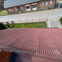 Widok z góry na falisty, bordowy dach z widocznymi elementami uszczelniającymi przy połączeniu z innym dachem, w tle budynek z czerwonej cegły, droga i zieleń.
