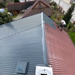 JKalpin Jan Kaźmierczak - Malowanie dachu z blachy trapezowej: widok z góry na dekarza zabezpieczonego linami, malującego czerwoną blachę na szaro, z widocznym fragmentem pomalowanej i niepomalowanej powierzchni dachu.