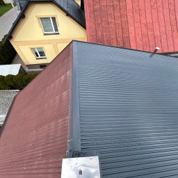 JKalpin Jan Kaźmierczak - Widok z góry na dwa dachy: jeden z blachy trapezowej w kolorze grafitowym, drugi z blachy w kolorze ceglastym; w tle widoczny fragment żółtego domu z ogrodem.