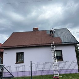 JKalpin Jan Kaźmierczak - Pracownik na drabinie, w trakcie malowania dachu z blachy trapezowej na szaro, widoczny fragment dachu w kolorze ceglastym, dom z jasną elewacją i numerem 27, pochmurne niebo.
