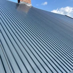 Szary dach z blachy trapezowej, widoczny komin z cegły i fragment błękitnego nieba z chmurami.