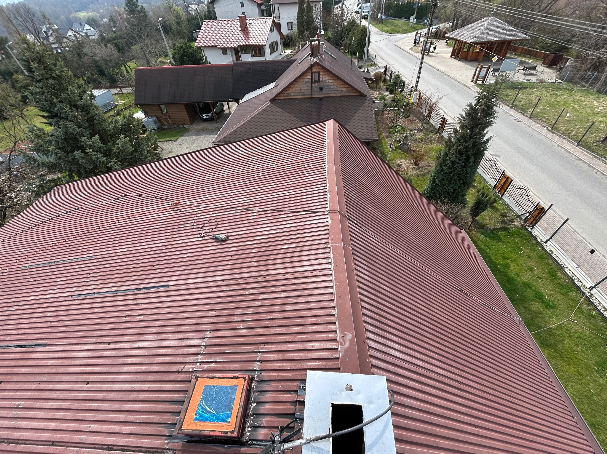 Widok z góry na czerwony, blaszany dach z widocznym świetlikiem zabezpieczonym folią, kominem i śladami napraw. W tle zabudowa jednorodzinna i droga.