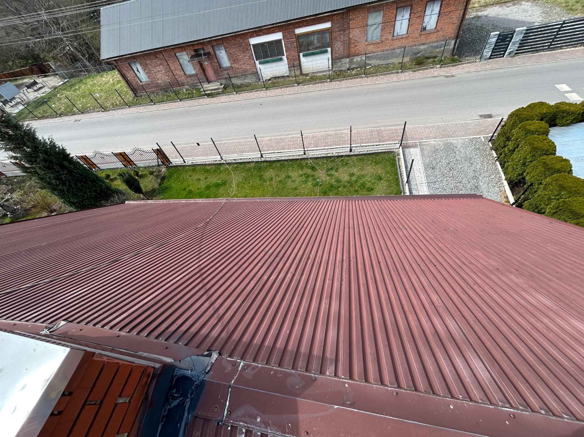 Widok z góry na falisty, bordowy dach z widocznymi elementami uszczelniającymi przy połączeniu z innym dachem, w tle budynek z czerwonej cegły, droga i zieleń.