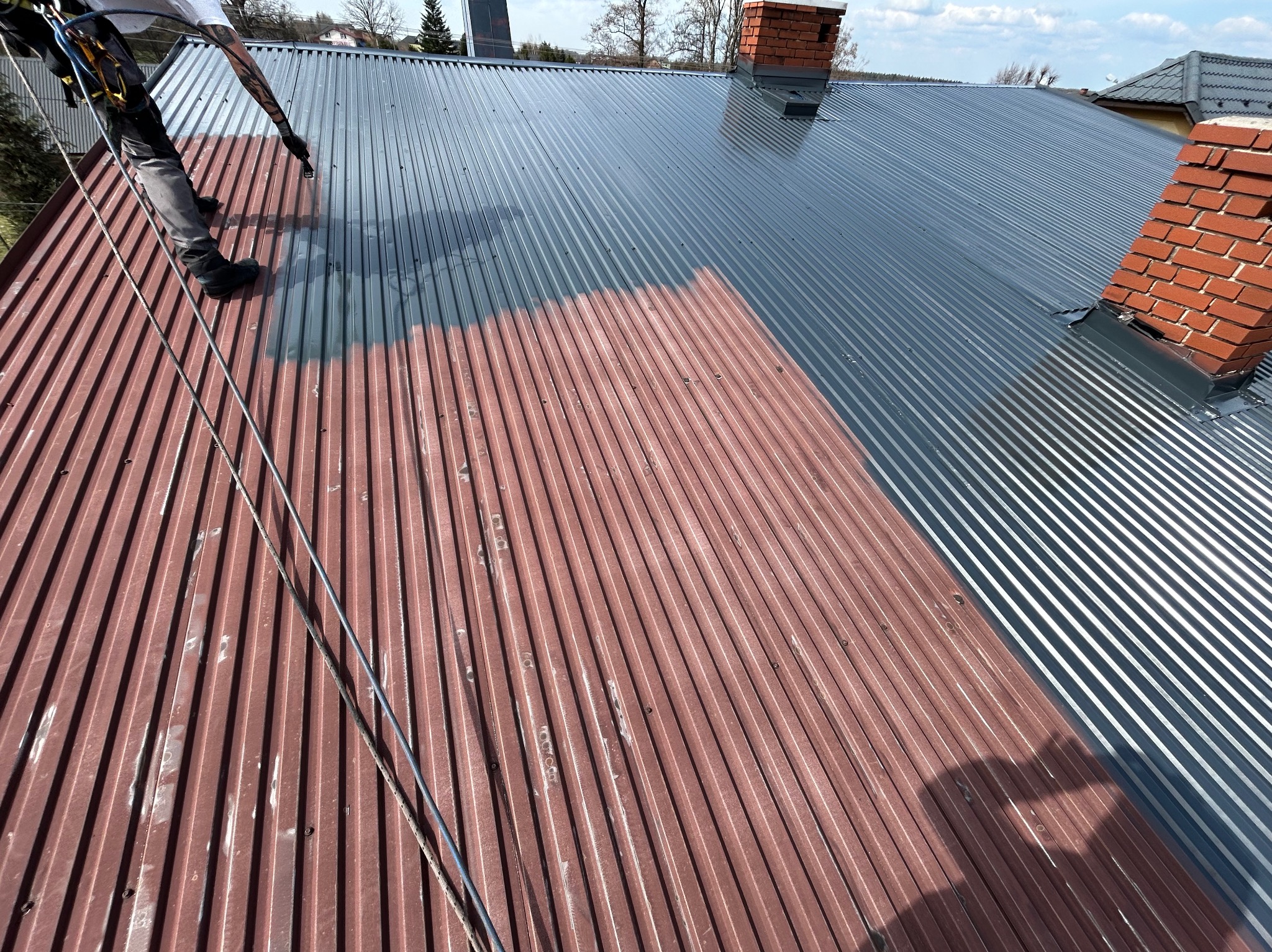 Pracownik zabezpieczony linami maluje blaszany dach na szaro, widoczny fragment dachu w kolorze ceglastym przed malowaniem, w tle komin z cegły i zabudowa.