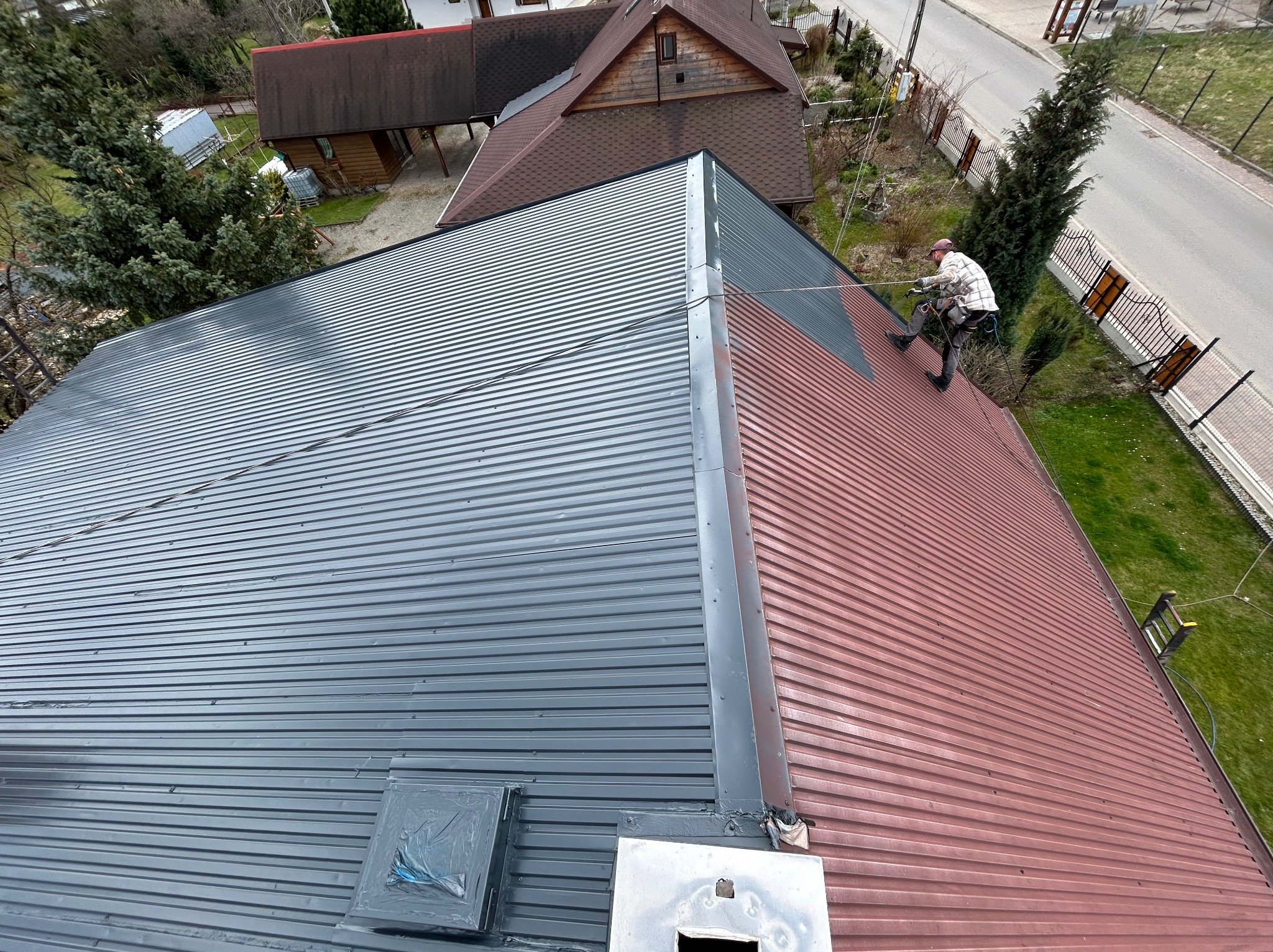 Malowanie dachu z blachy trapezowej: widok z góry na dekarza zabezpieczonego linami, malującego czerwoną blachę na szaro, z widocznym fragmentem pomalowanej i niepomalowanej powierzchni dachu.