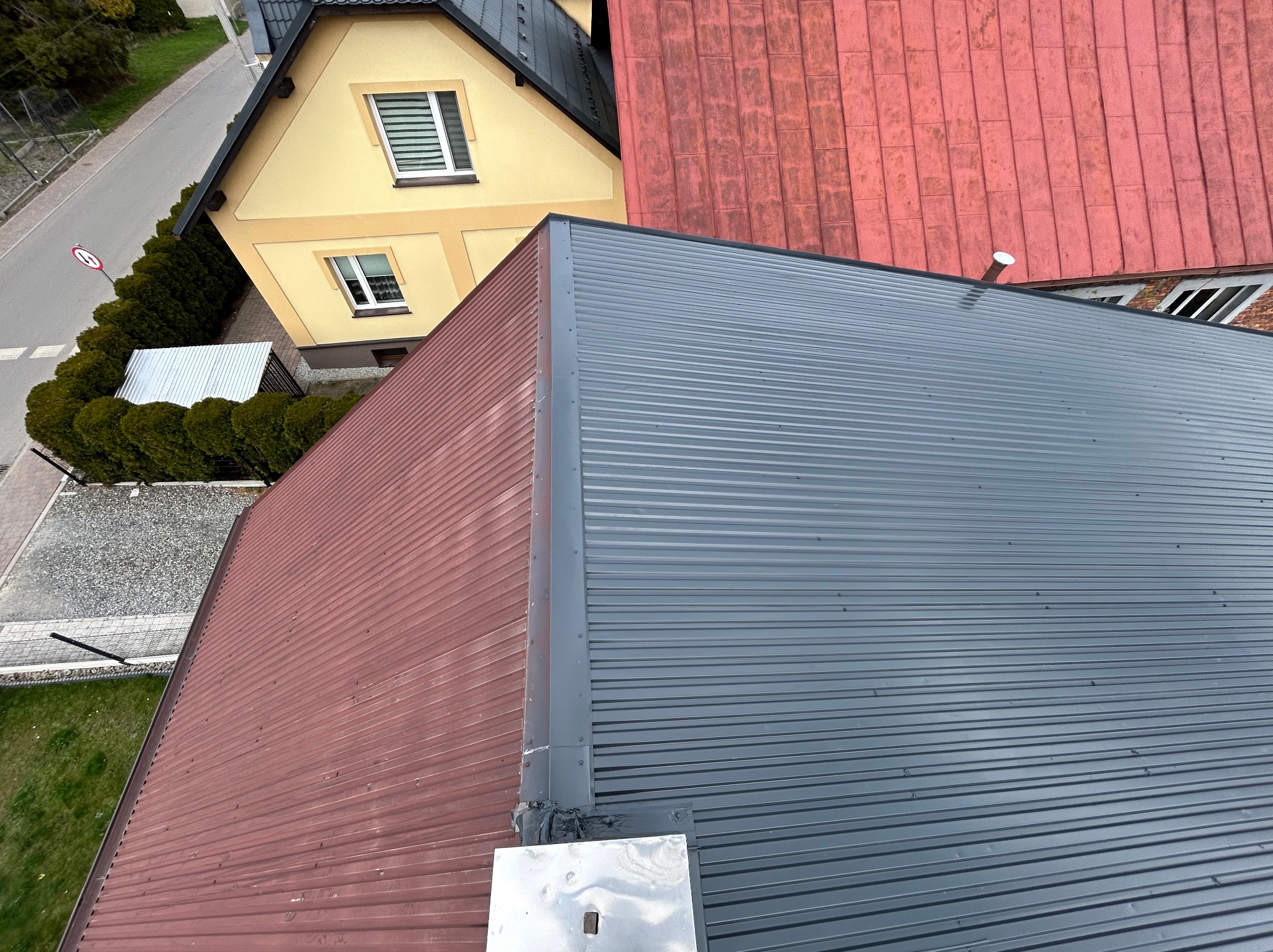 Widok z góry na dwa dachy: jeden z blachy trapezowej w kolorze grafitowym, drugi z blachy w kolorze ceglastym; w tle widoczny fragment żółtego domu z ogrodem.