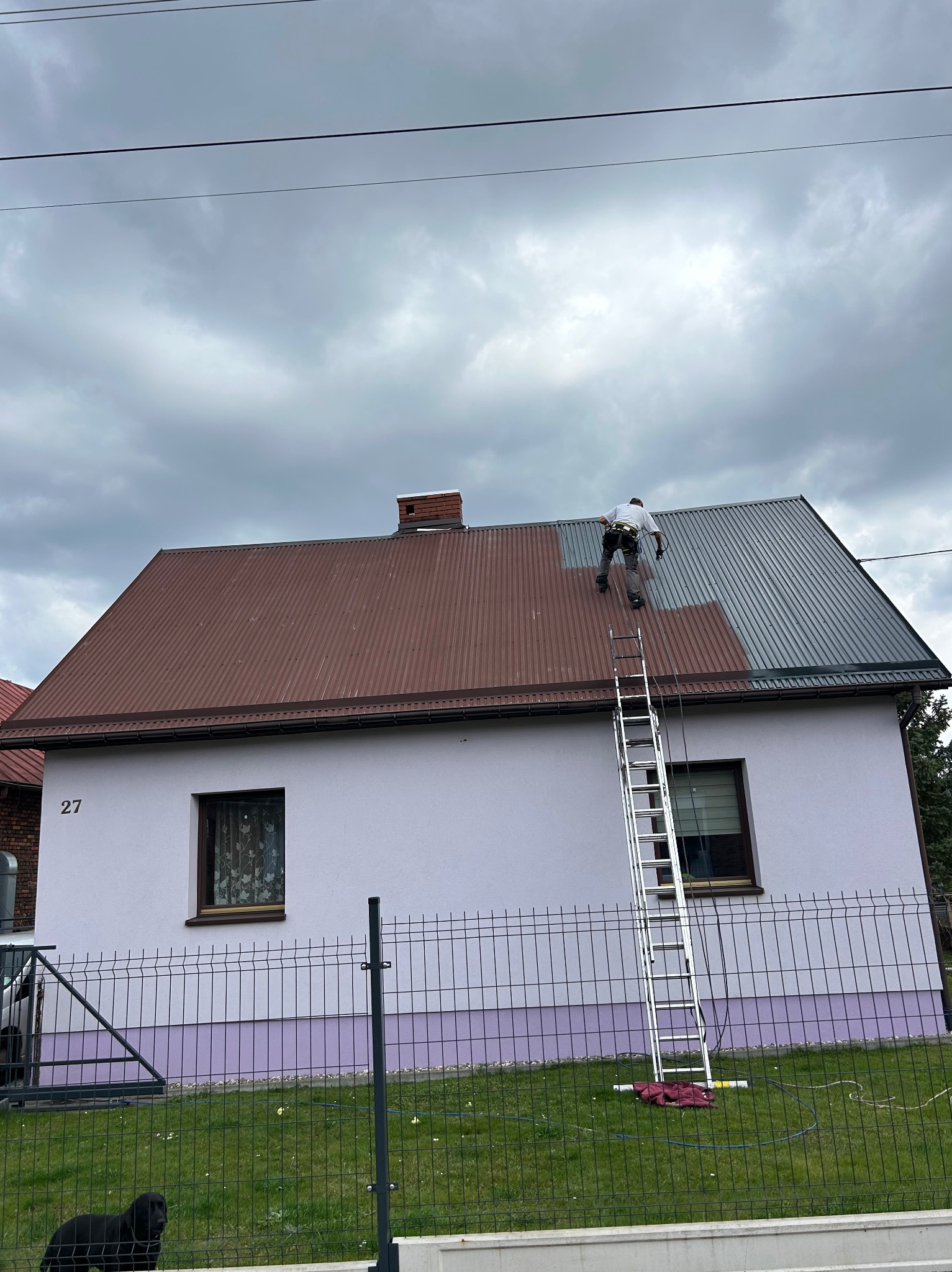 Pracownik na drabinie, w trakcie malowania dachu z blachy trapezowej na szaro, widoczny fragment dachu w kolorze ceglastym, dom z jasną elewacją i numerem 27, pochmurne niebo.