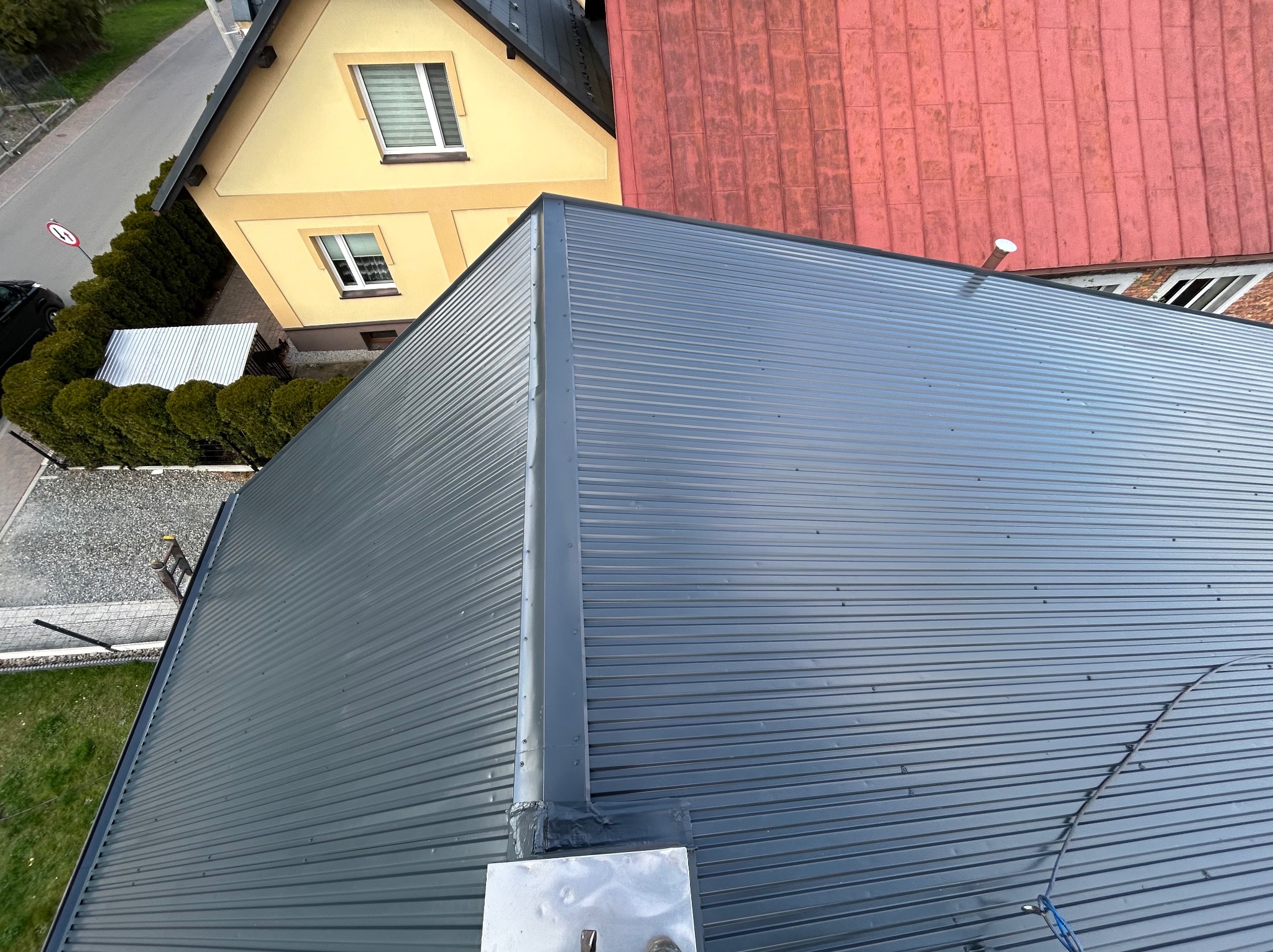 Szary dach z blachy trapezowej widziany z góry, z widocznymi elementami mocującymi, fragmentami sąsiednich budynków z dachami w kolorze czerwonym i żółtym oraz zielenią w tle.