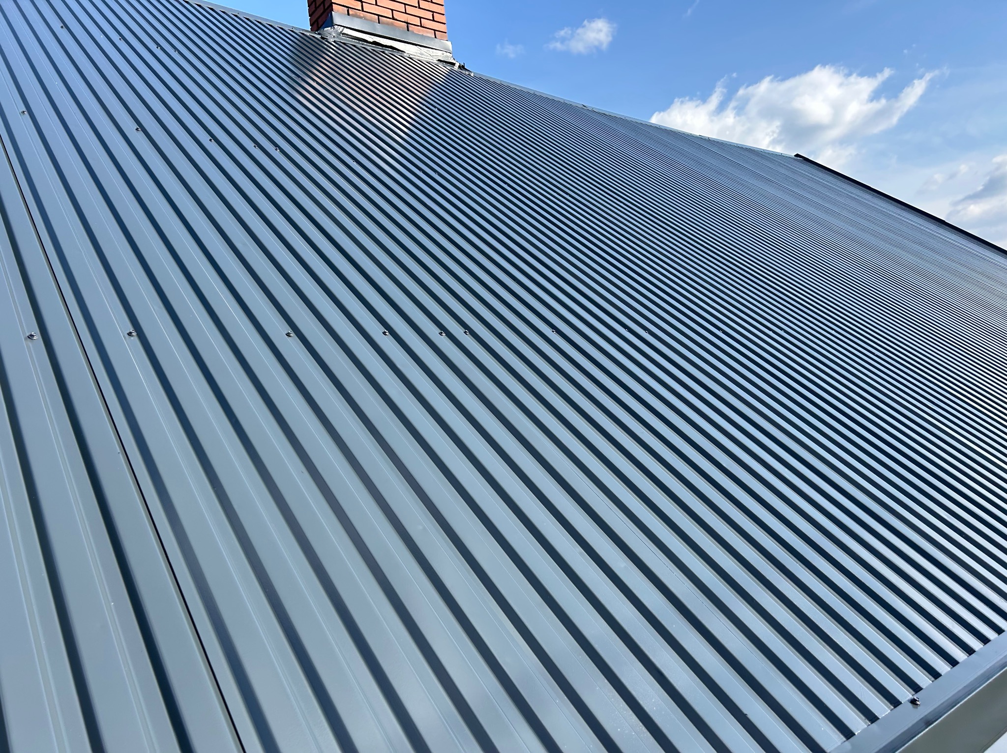 Szary dach z blachy trapezowej, widoczny komin z cegły i fragment błękitnego nieba z chmurami.