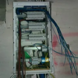 Otwarta skrzynka rozdzielcza z widocznymi bezpiecznikami, przewodami w różnych kolorach i listwami zaciskowymi, zamontowana na surowej ścianie.