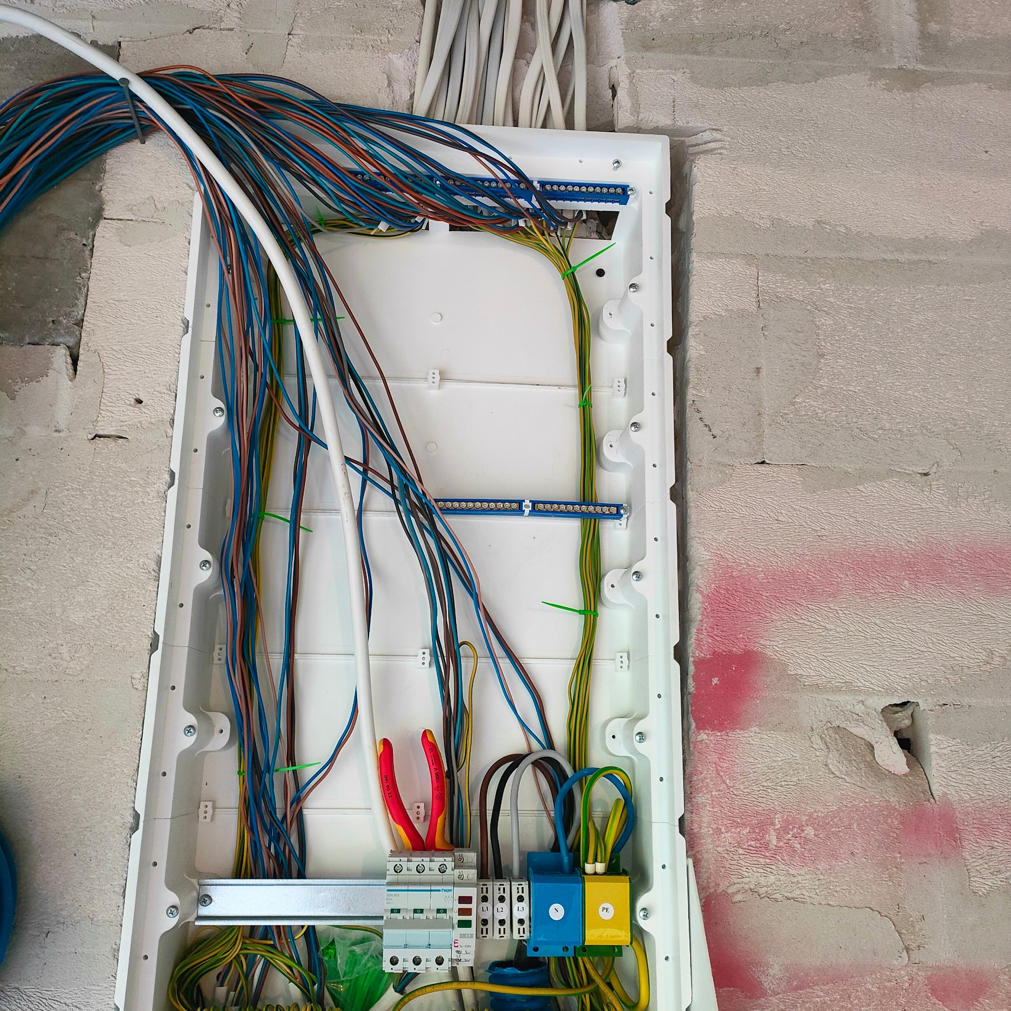 Otwarta skrzynka elektryczna w surowym stanie, widoczne liczne przewody w różnych kolorach, listwa zaciskowa, bezpieczniki, oraz narzędzia - czerwone szczypce do zdejmowania izolacji, ściana w tle...