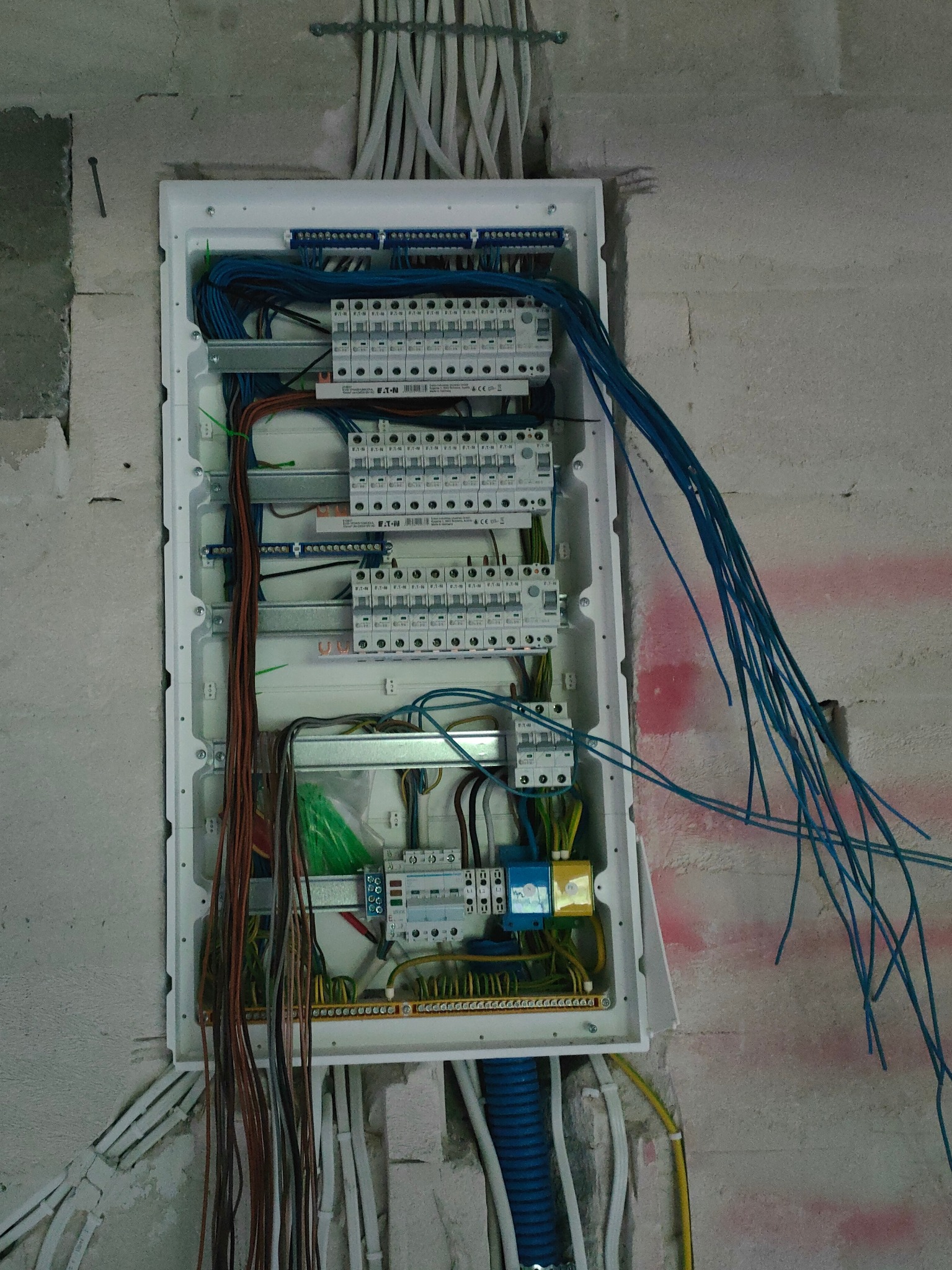 Otwarta skrzynka rozdzielcza z widocznymi bezpiecznikami, przewodami w różnych kolorach i listwami zaciskowymi, zamontowana na surowej ścianie.