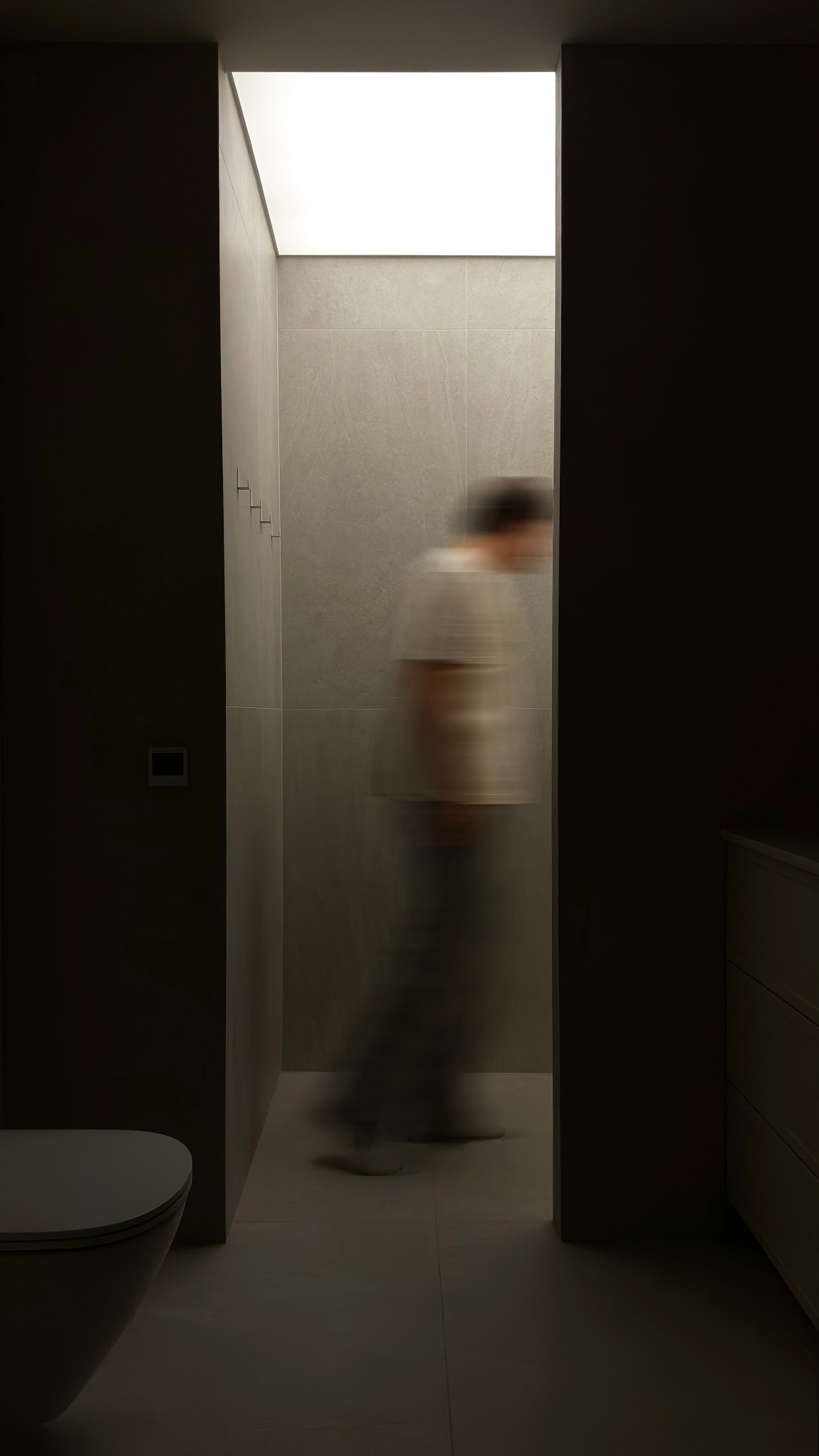 Minimalistyczna łazienka z szarymi płytkami, podłużnym oknem w suficie wpuszczającym światło i rozmytą sylwetką osoby przechodzącej przez kadr, widok z perspektywy toalety.