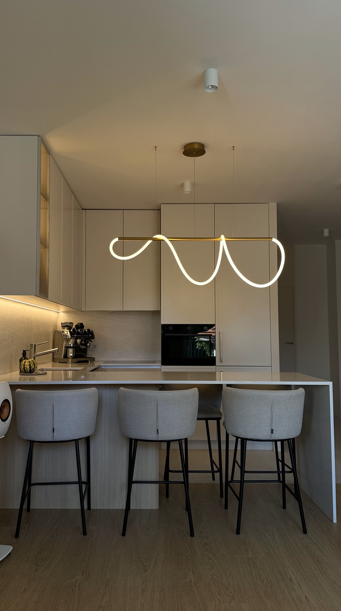 Nowoczesna kuchnia z wyspą, oświetlona podłużną lampą LED z falistym wzorem, trzy szare hokery z czarnymi nogami ustawione przy blacie.