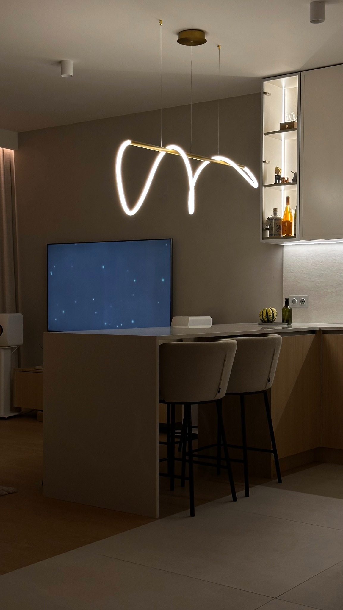 Nowoczesna kuchnia z wyspą, krzesłami barowymi i designerską lampą LED w kształcie abstrakcyjnej linii.