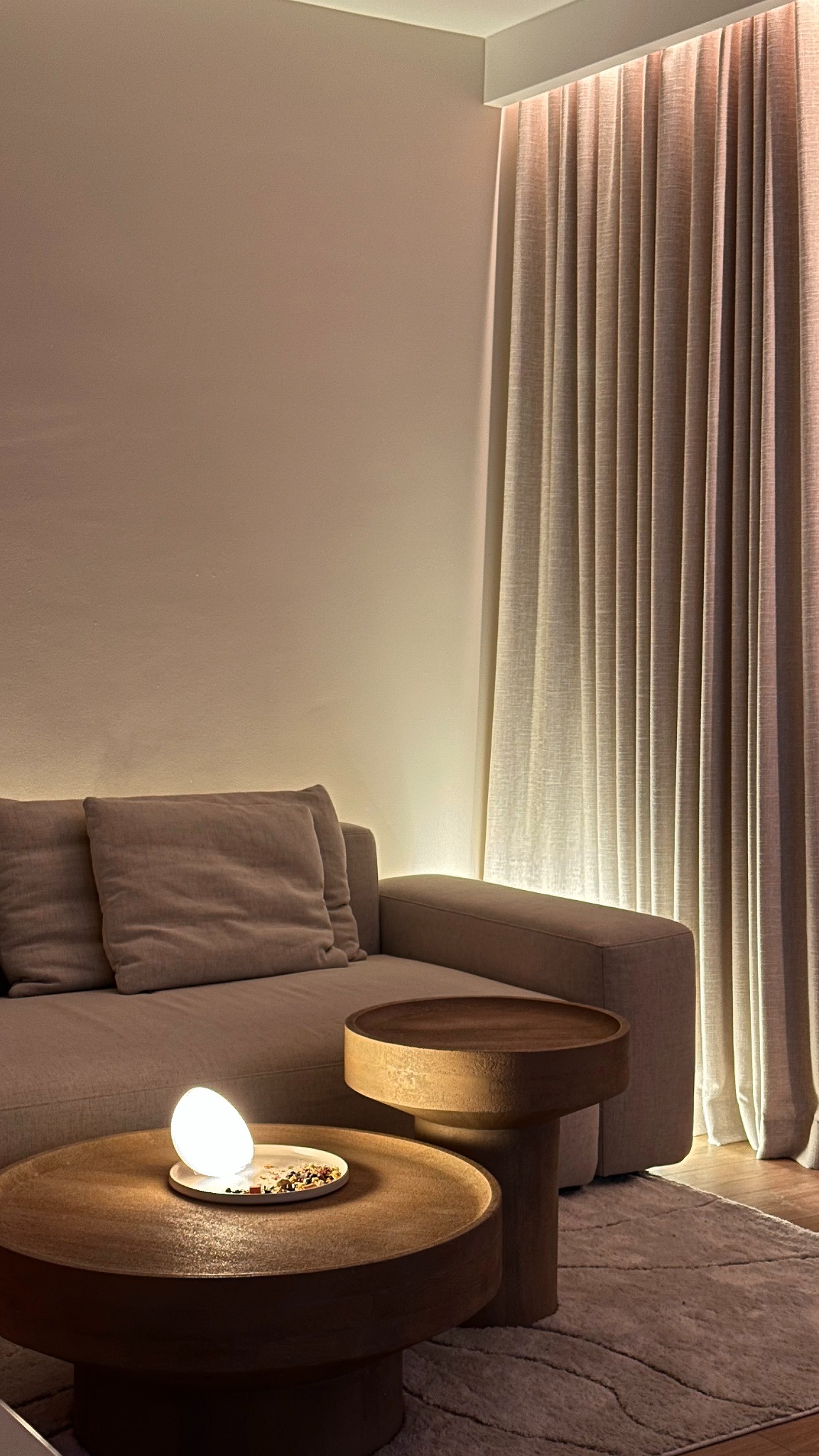 Minimalistyczny salon z beżową sofą, okrągłymi stolikami kawowymi z drewna i zasłonami podświetlonymi od góry, tworzącymi ciepłą atmosferę.