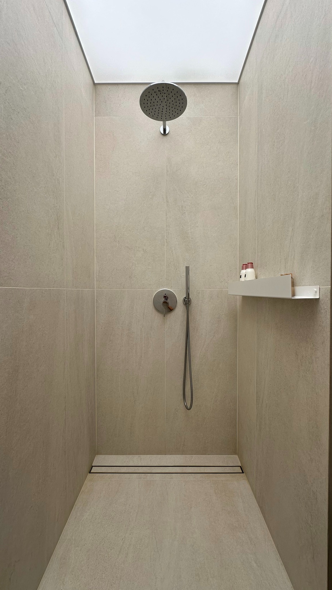 Minimalistyczna kabina prysznicowa z beżowymi płytkami, deszczownicą, półką z kosmetykami i odpływem liniowym.