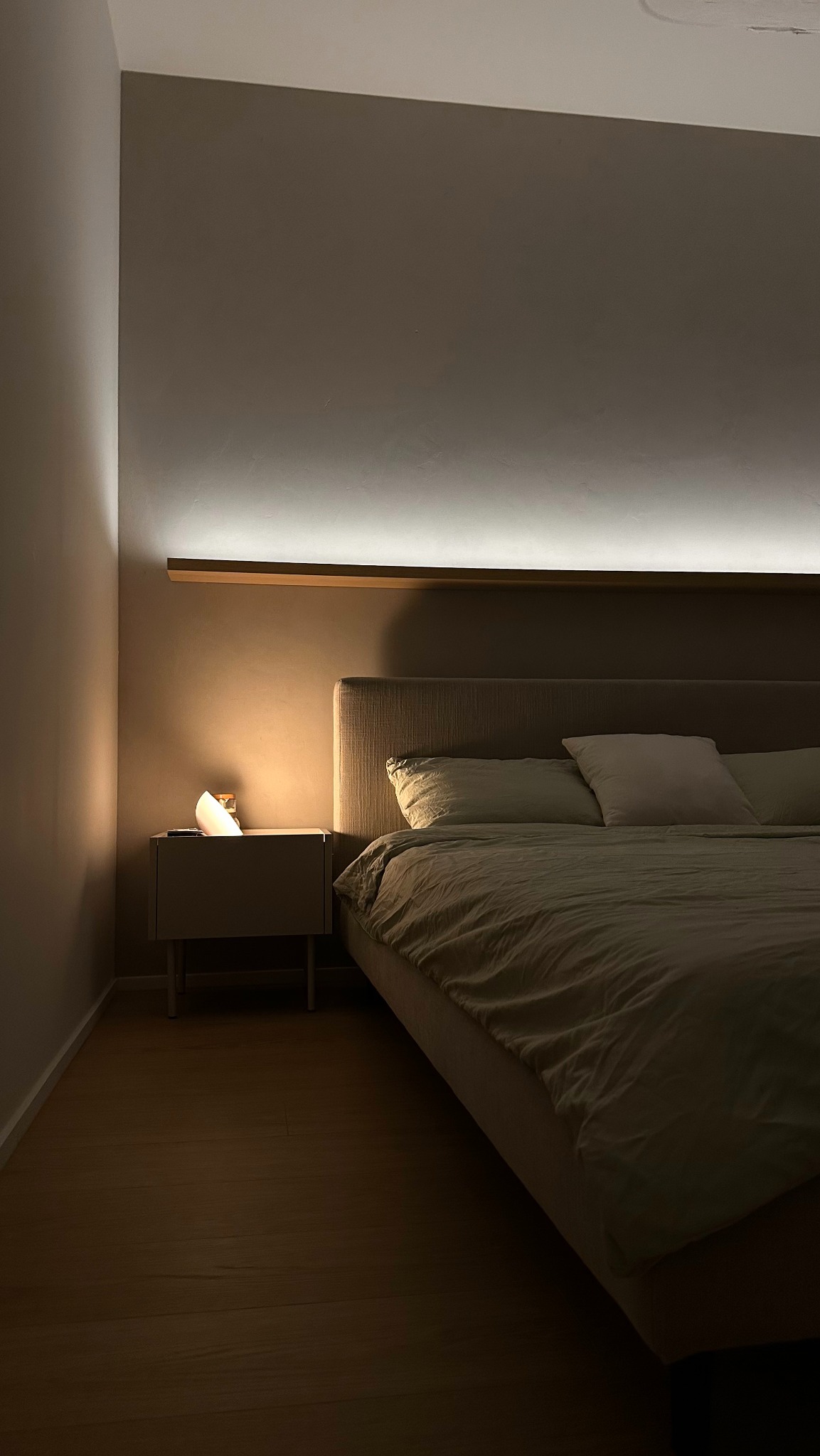 Minimalistyczna sypialnia z łóżkiem o szarej tapicerce, drewnianą półką nad wezgłowiem i podświetleniem LED, nocna szafka z nowoczesną lampką.