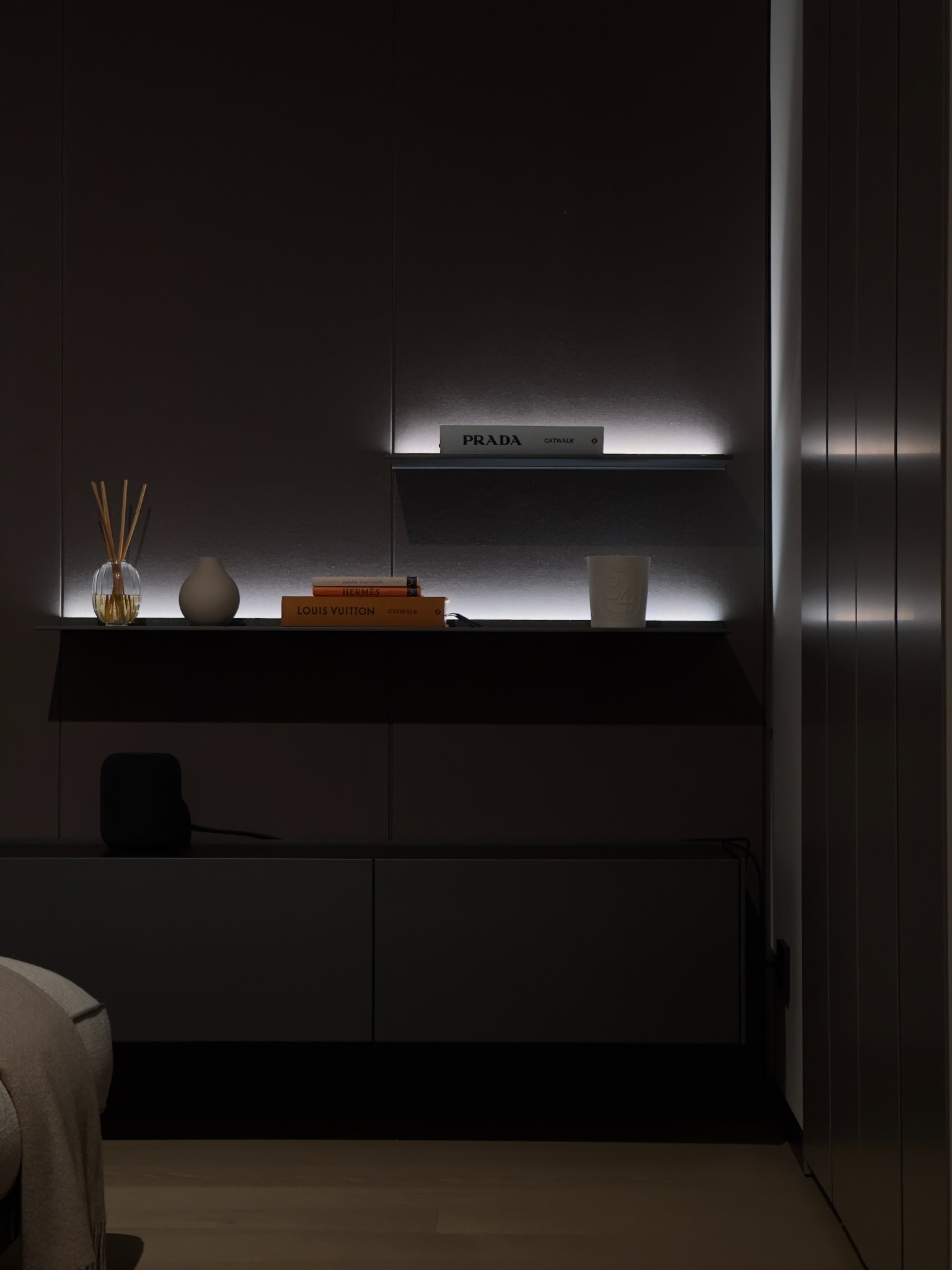 Stylowa, ciemna szafka z podświetlanymi półkami, na których znajdują się książki Prada i Louis Vuitton, dyfuzor zapachowy i ceramiczny wazon.
