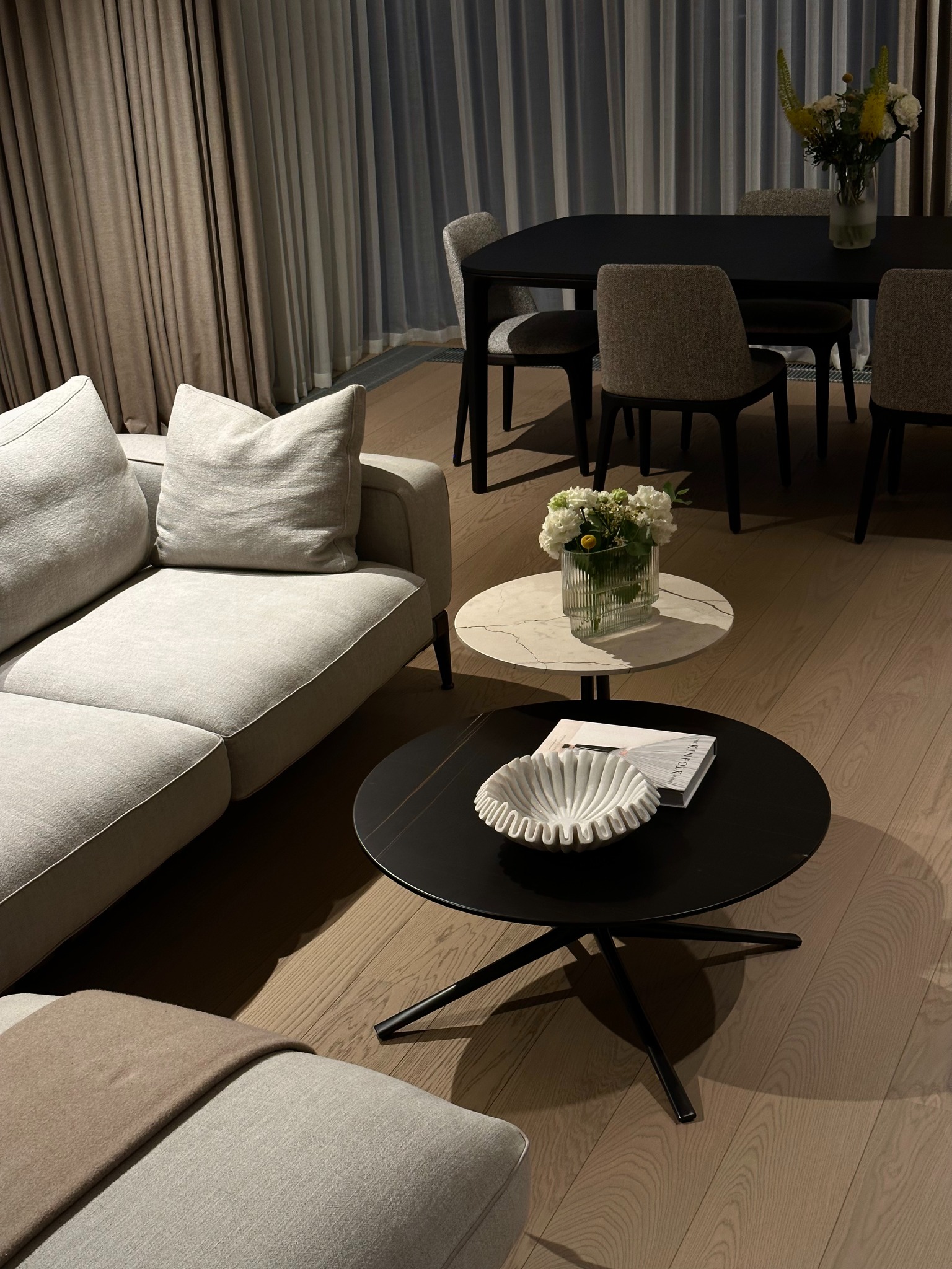 Jasnoszara sofa modułowa z beżowym pledem, okrągły stolik kawowy z czarnym blatem, biała dekoracyjna misa i książka, stolik pomocniczy z marmurowym blatem i wazonem z białymi kwiatami, stół jadalny...