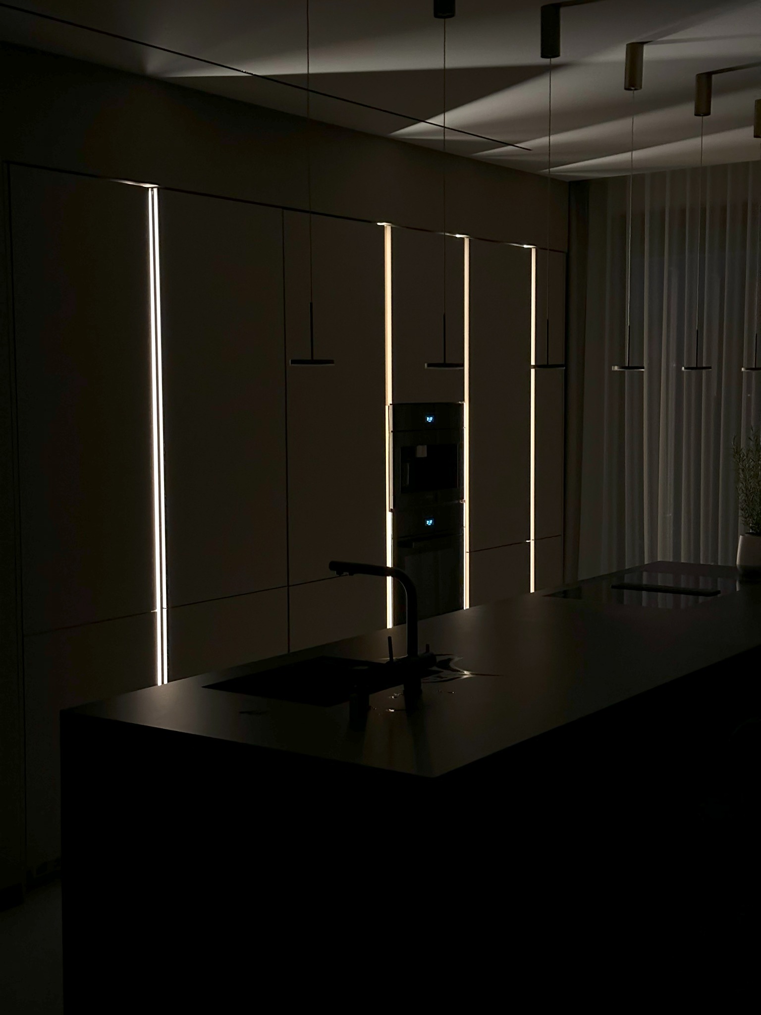 Nowoczesna kuchnia z ciemnymi szafkami oświetlonymi pionowymi paskami LED, czarny blat wyspy kuchennej z minimalistyczną baterią, wiszące lampy i delikatne zasłony w tle.
