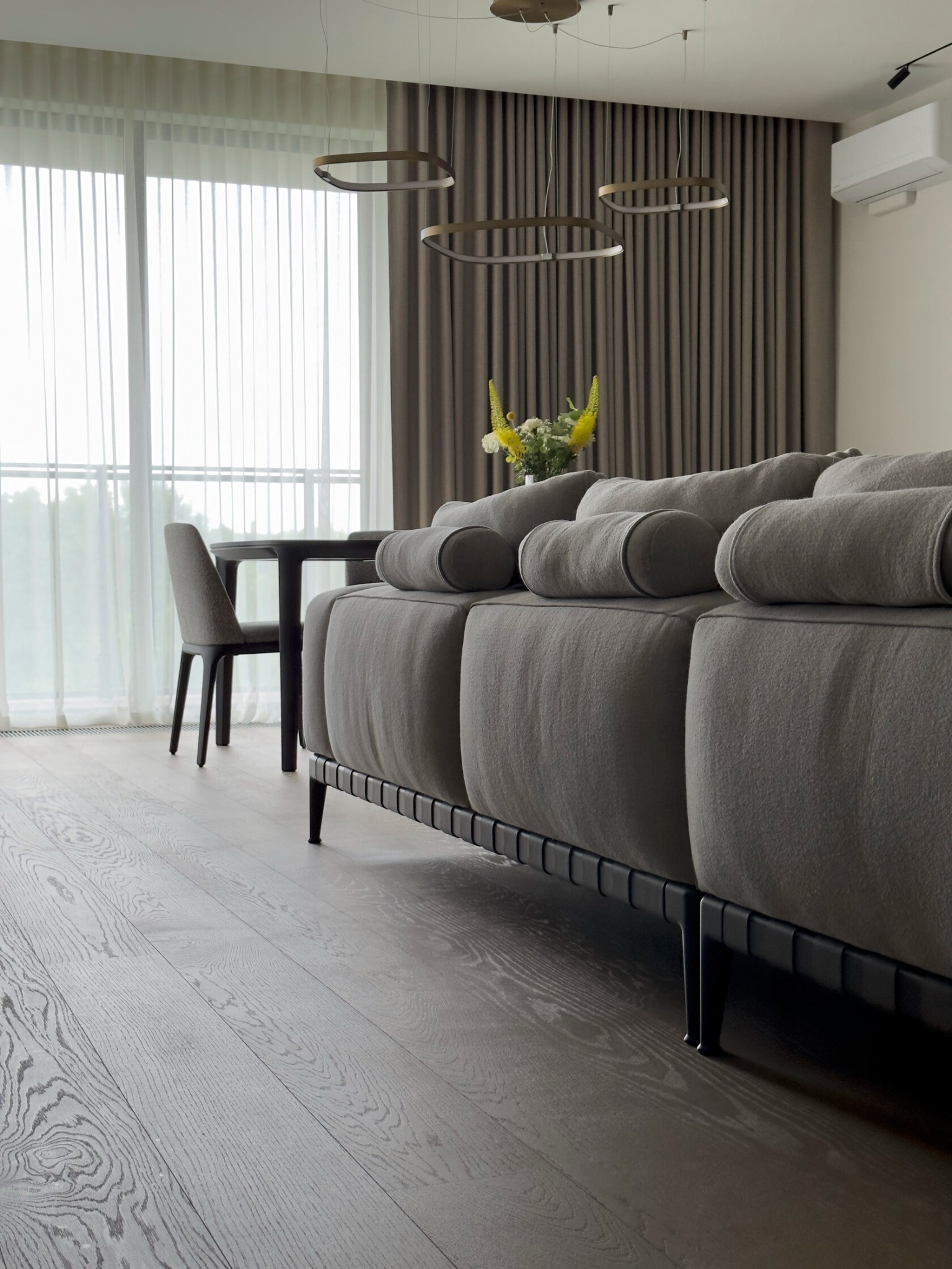 Nowoczesny salon z szarym, modułowym narożnikiem, ciemną drewnianą podłogą, okrągłym stołem i krzesłami w tle, oświetlony wiszącymi lampami w kształcie obręczy i naturalnym światłem z okna z firanami.