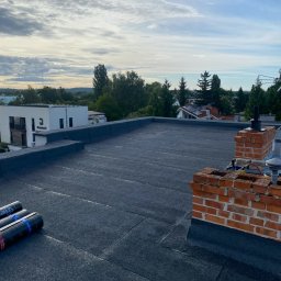 Roofbud - Płaski dach pokryty papą termozgrzewalną, widoczne kominy z cegły i rolki papy. Tło stanowi zabudowa jednorodzinna i błękitne niebo z chmurami.