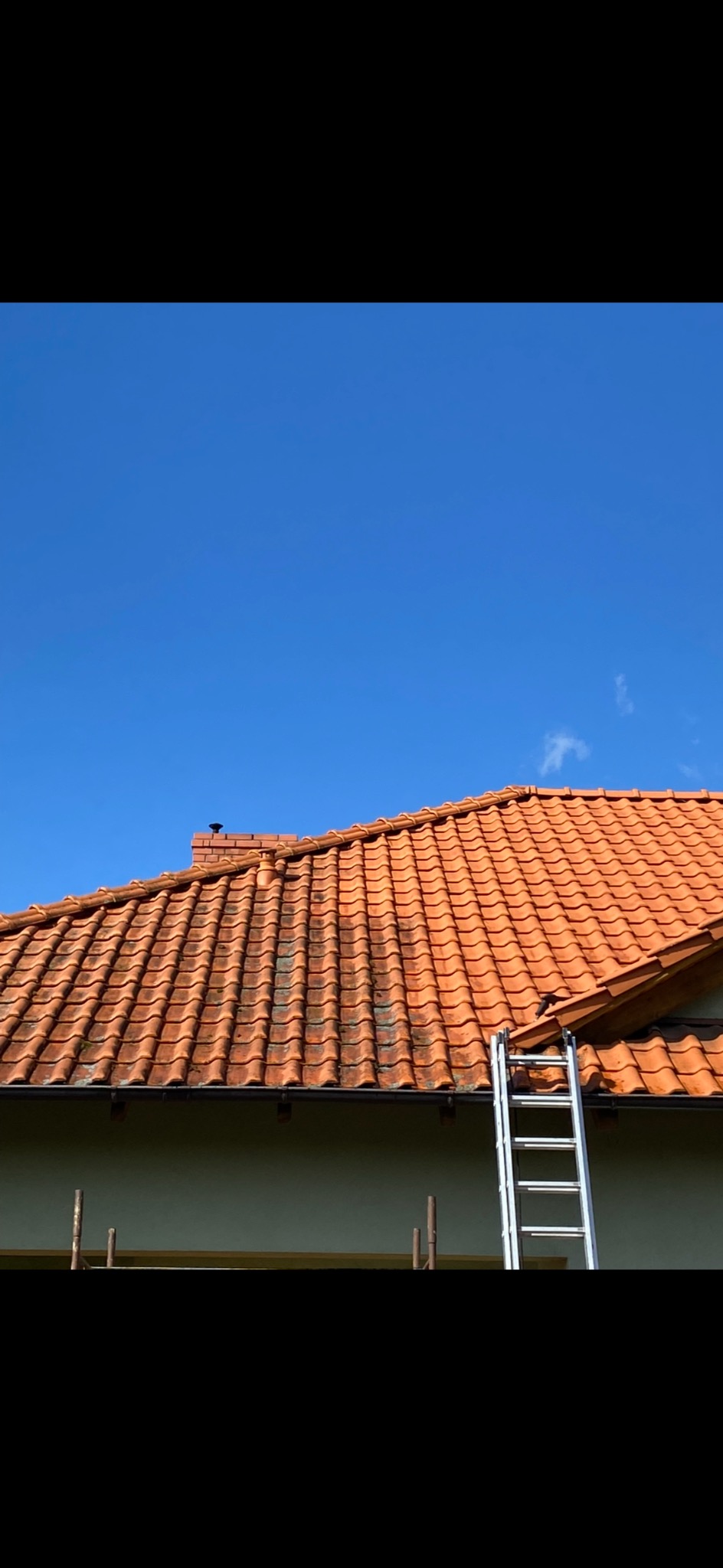 Ceglasty dach z kominem i aluminiową drabiną opartą o rynnę na tle błękitnego nieba. Widoczne ślady zabrudzeń na dachówkach.