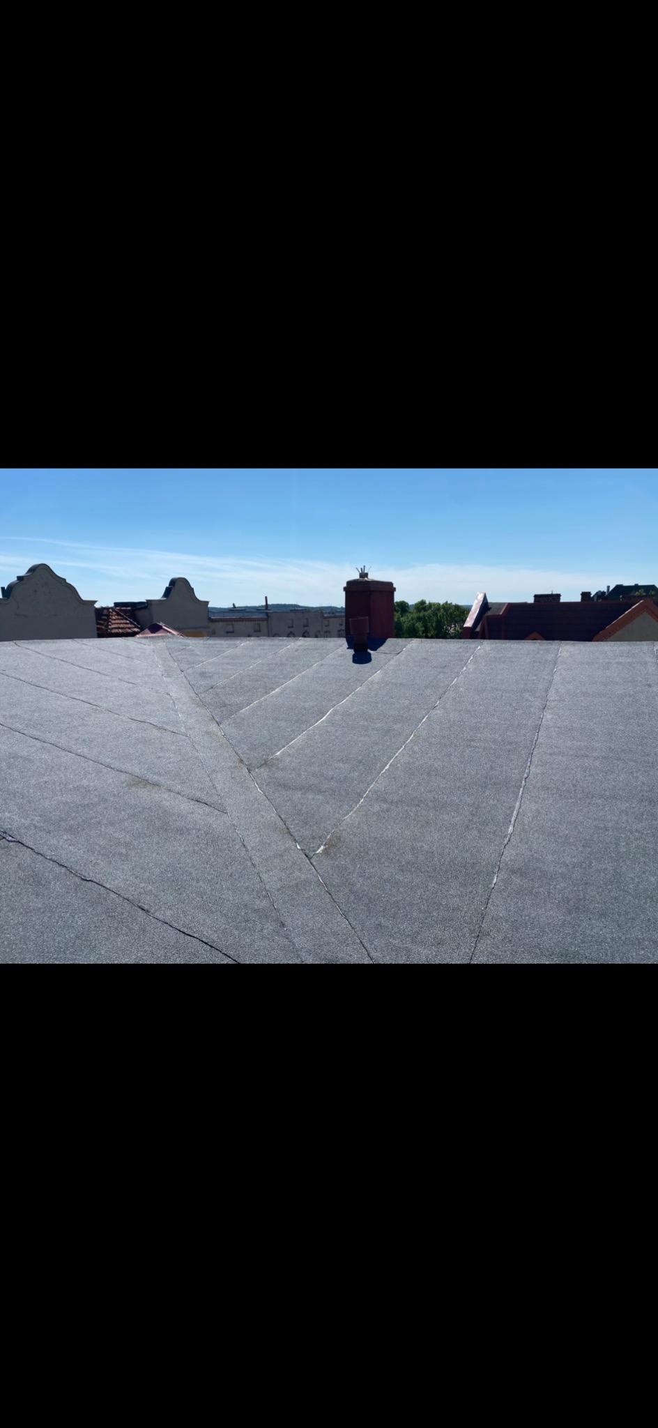 Szary, płaski dach pokryty papą na tle błękitnego nieba i zabudowań miejskich. Widoczny komin i inne dachy w oddali. Układ połaci dachu tworzy geometryczny wzór.