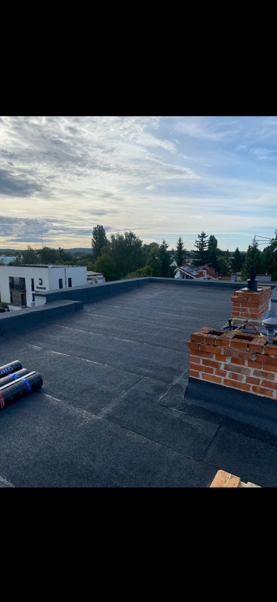 Płaski dach pokryty papą termozgrzewalną, widoczne kominy z cegły i rolki papy. Tło stanowi zabudowa jednorodzinna i błękitne niebo z chmurami.