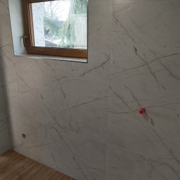 WLAD-BUD - Ściana łazienki wyłożona białymi płytkami imitującymi marmur, z oknem w drewnianej ramie. Widoczne fragmenty podłogi z paneli i grzejnika.